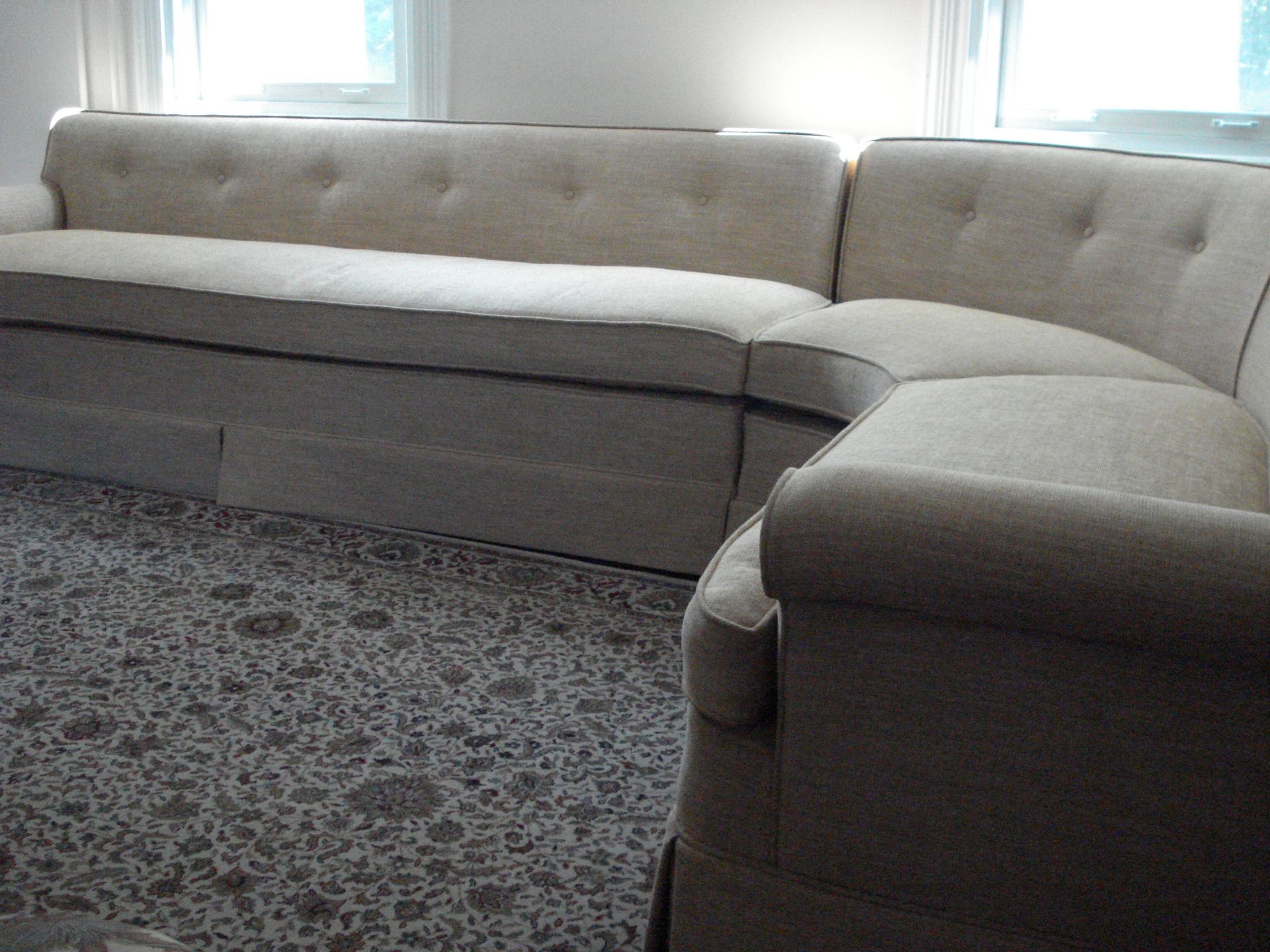 Beige sectional sofa with dark trim in front of windows, on patterned carpet.