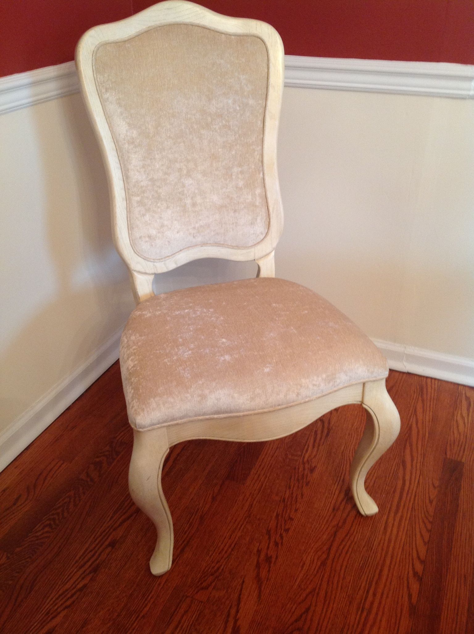 Cream-colored velvet chair in a corner, with ornate curved legs and back against a red wall and wood floor.