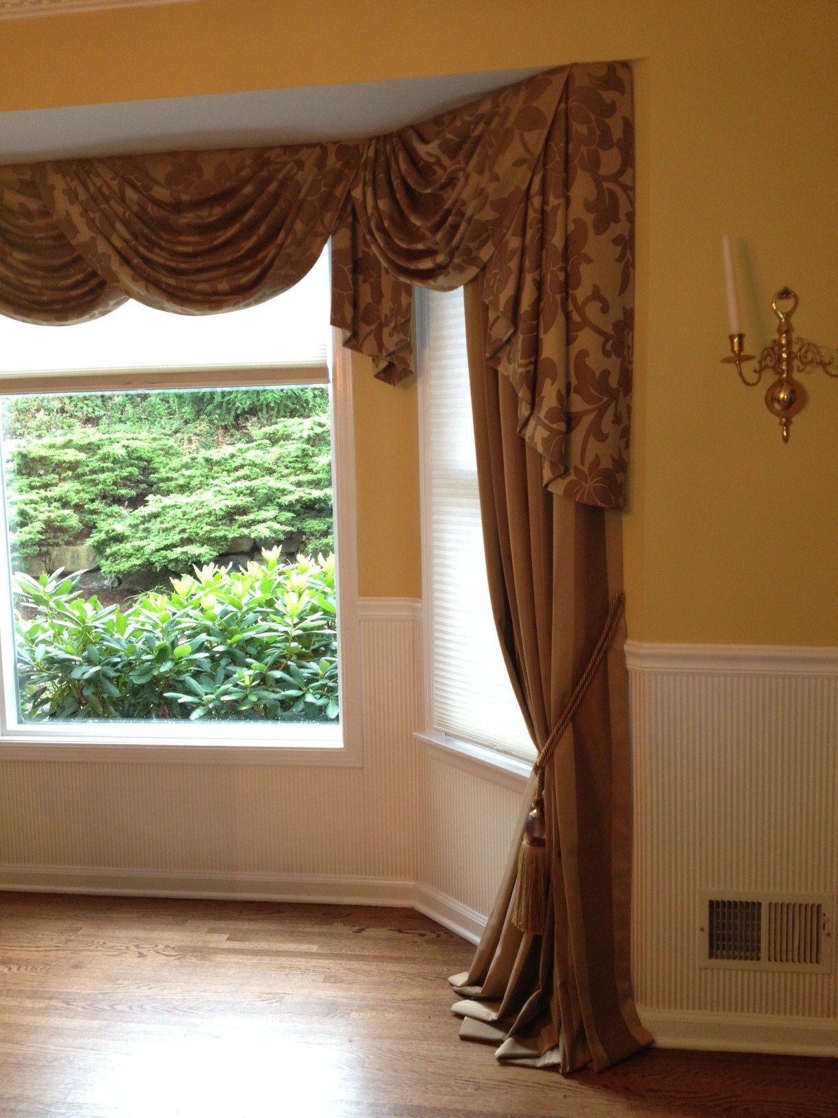 A bay window with tan drapes and a valance, a view of green foliage outside, and a sconce on a yellow wall.