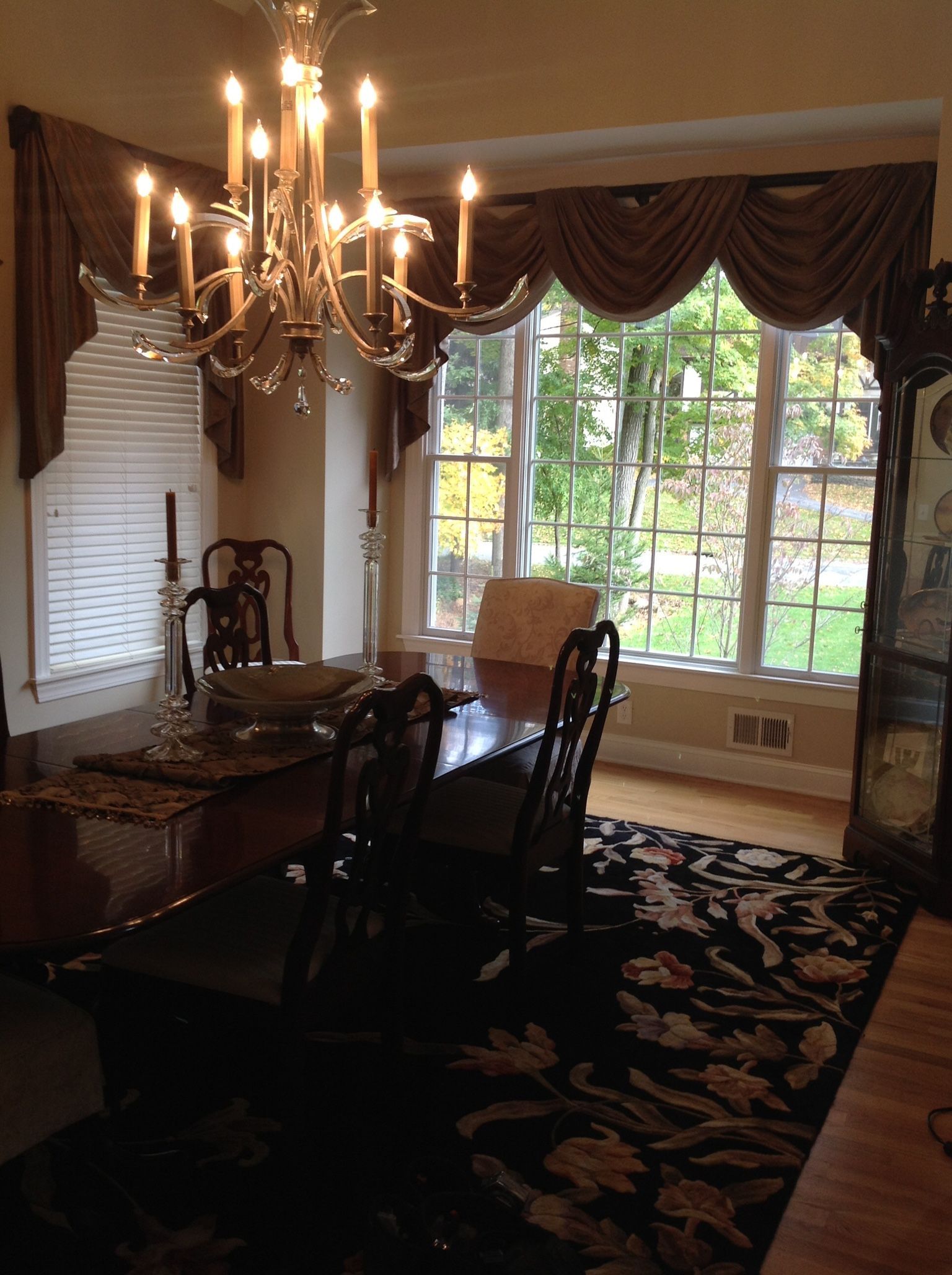 Formal dining room: dark table, chairs, chandelier, patterned rug, and window with draped valance.