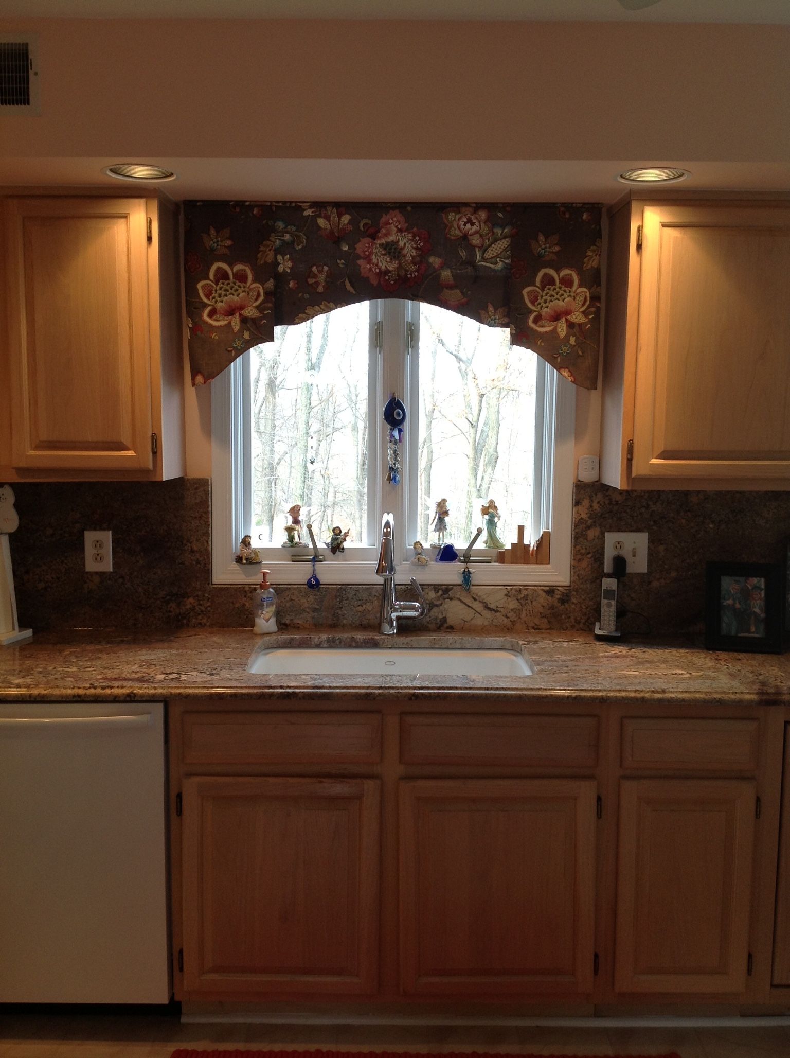 Kitchen sink with floral window valance above window, cabinets and countertops.