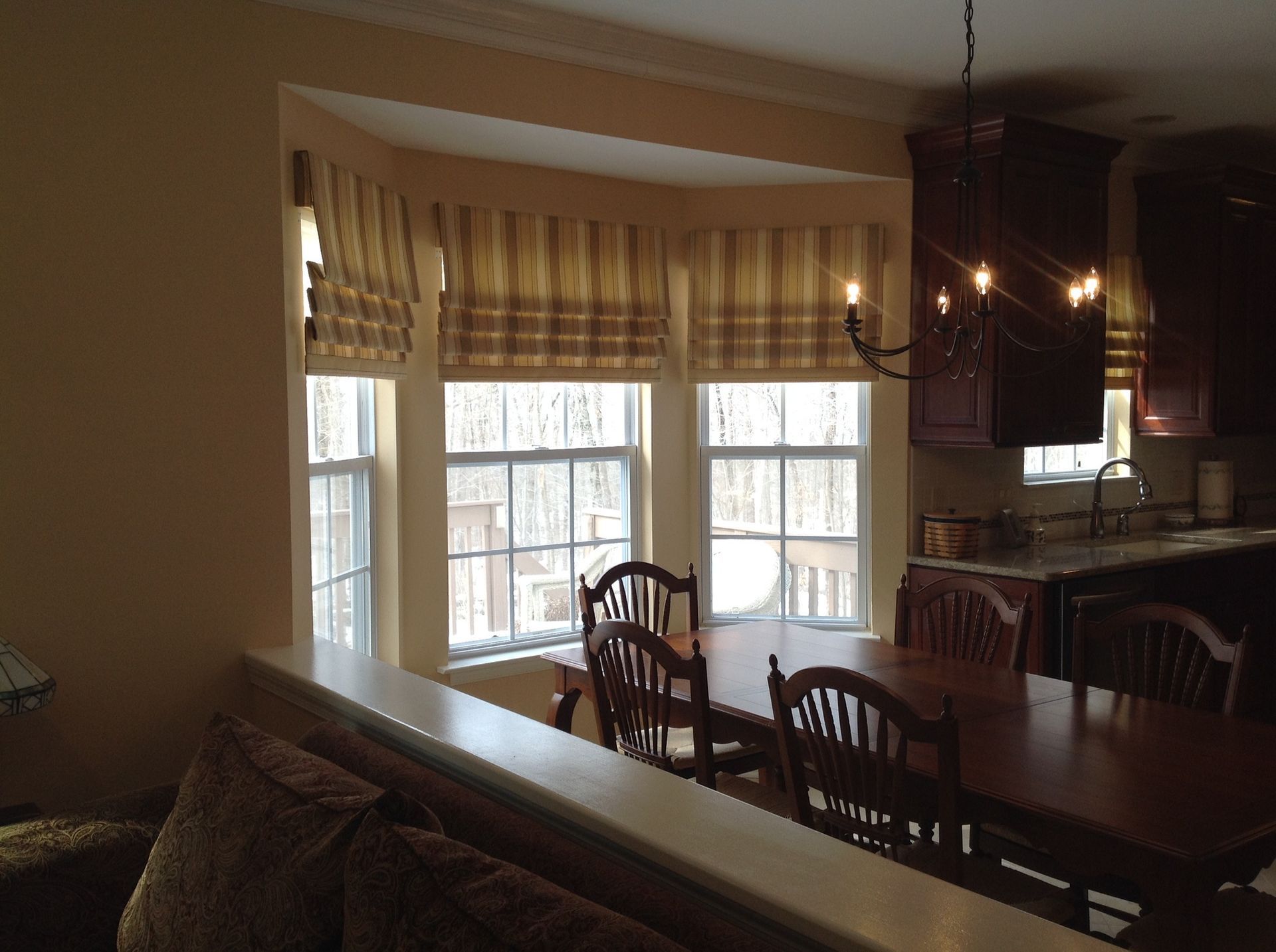 Dining room with bay window, table, chairs, and kitchen with dark cabinets. Neutral tones and warm light.