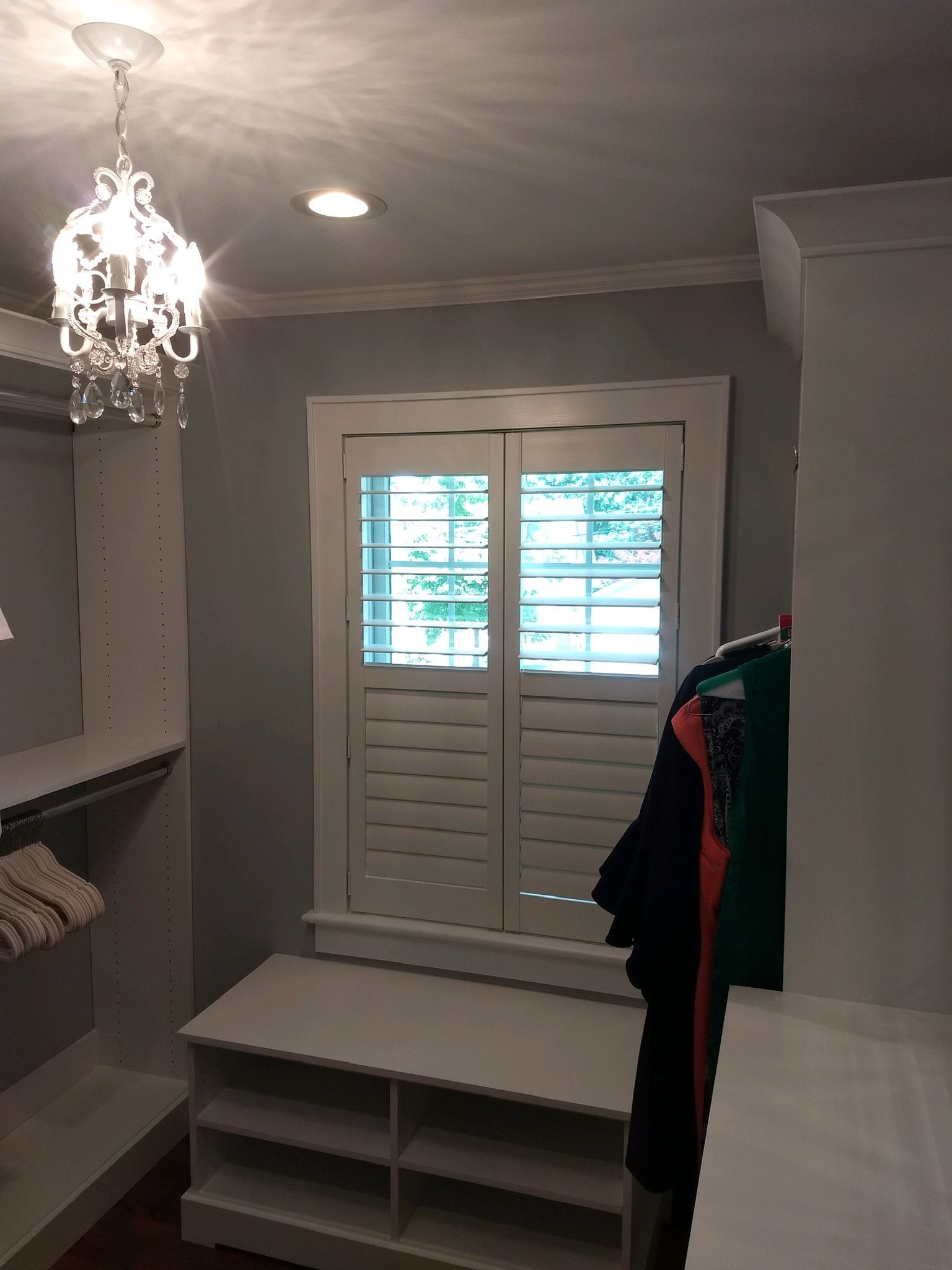 Walk-in closet with white shelves, shoe storage, and shutters; a crystal chandelier hangs overhead.