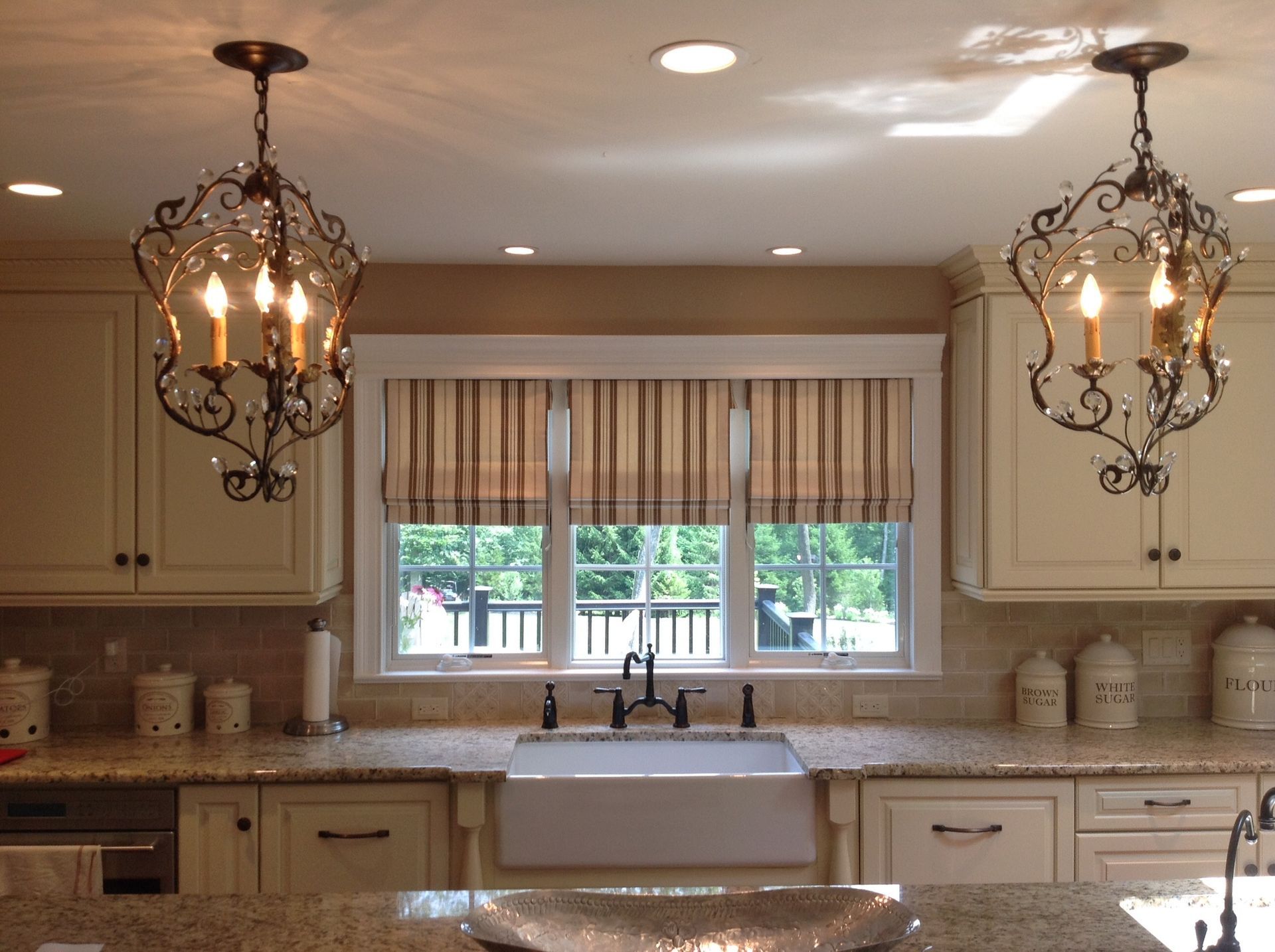 Kitchen with two ornate chandeliers, cream cabinets, striped window shades, and a farmhouse sink.
