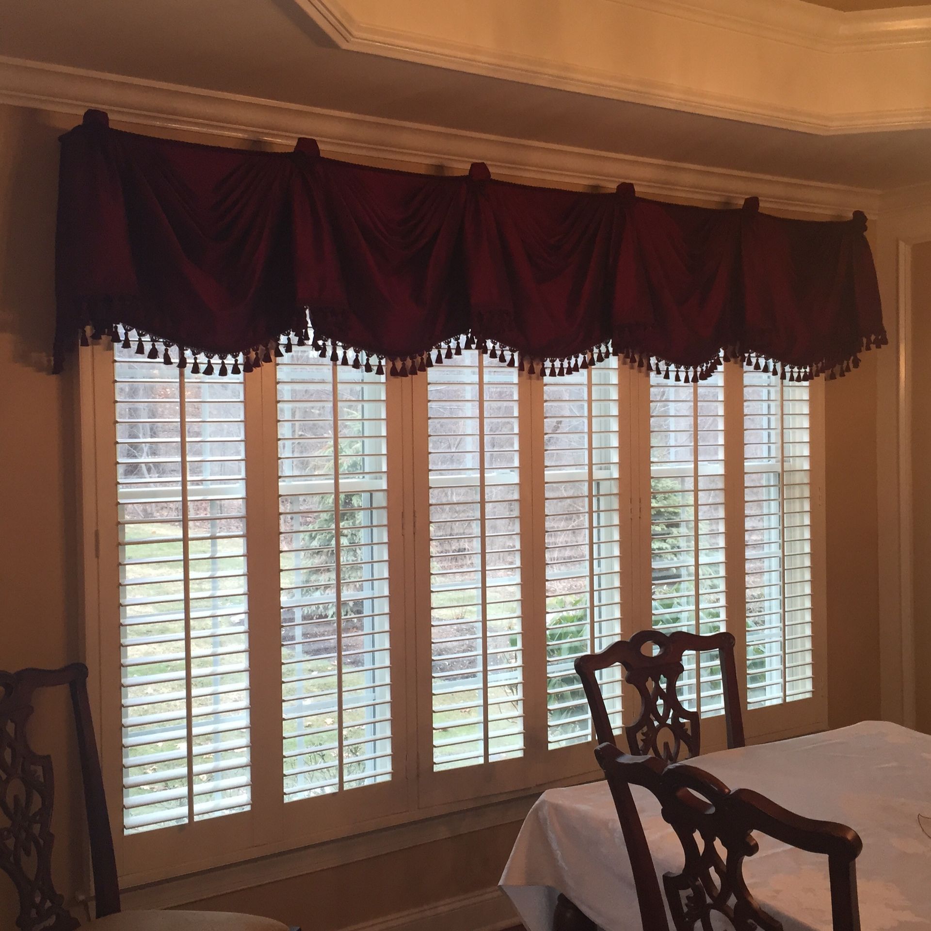 Maroon window valance with tassel trim over white shutters. Dining room setting.