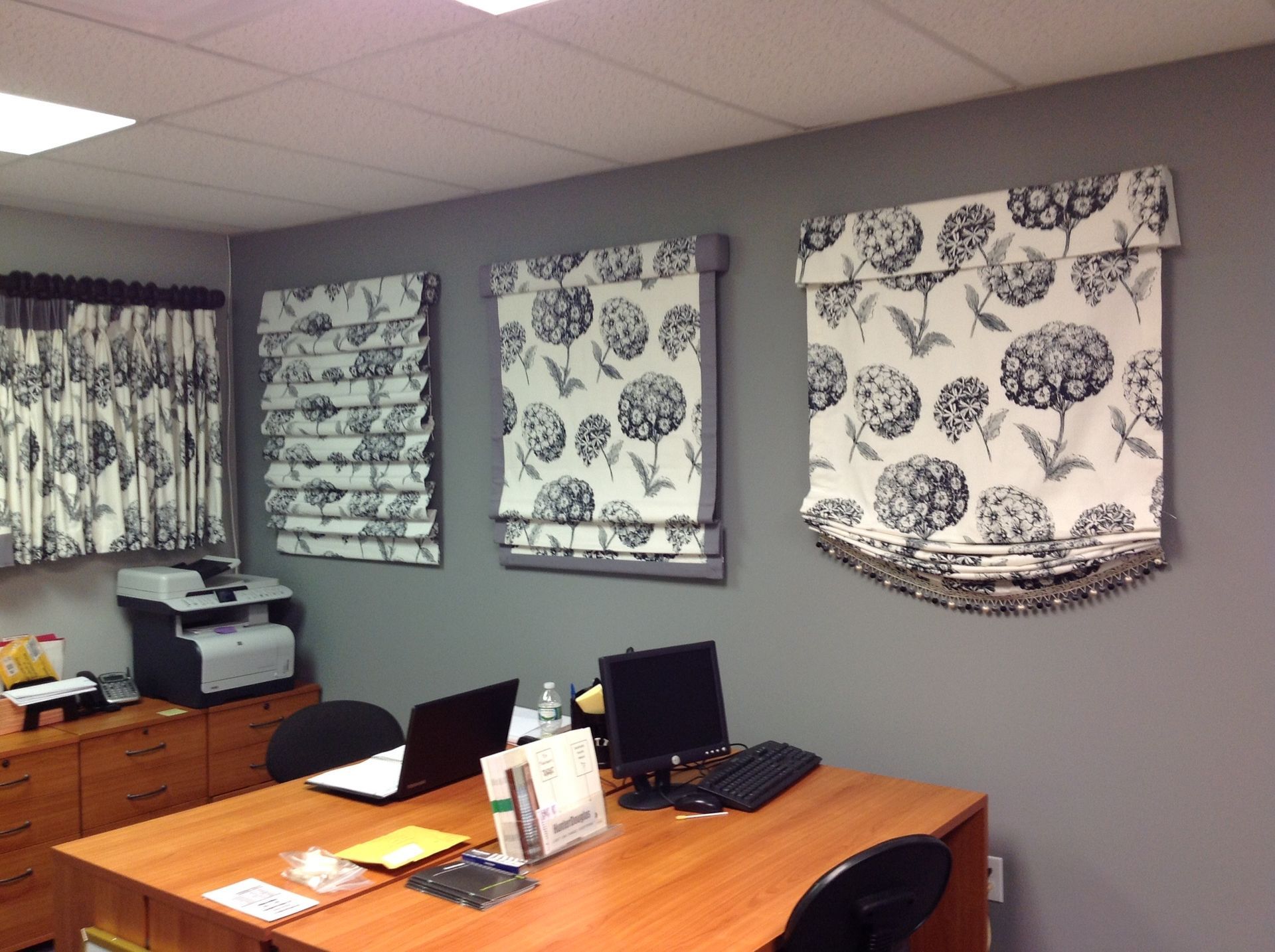 Office with desk, computer, and gray walls. Displaying various patterned window shades.