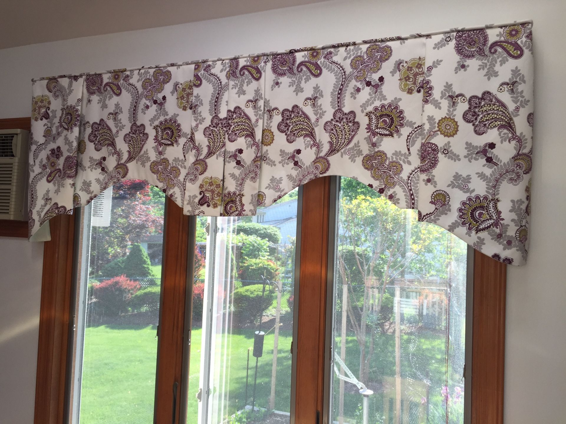 Window with floral valance; garden view. The valance is white with purple and yellow flowers.