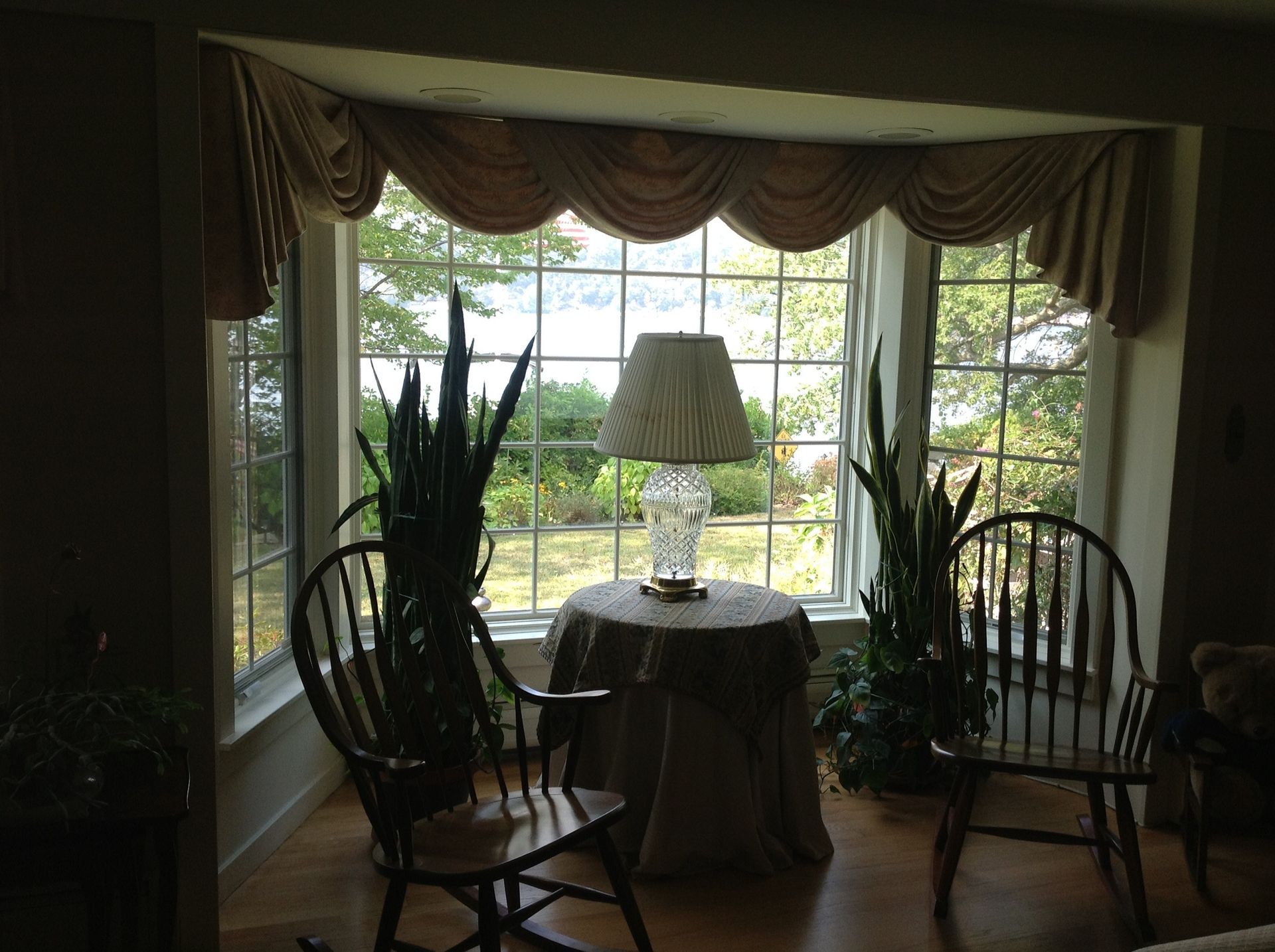 A window with plants, a lamp, and chairs, under a decorative valance, with an outdoor view.