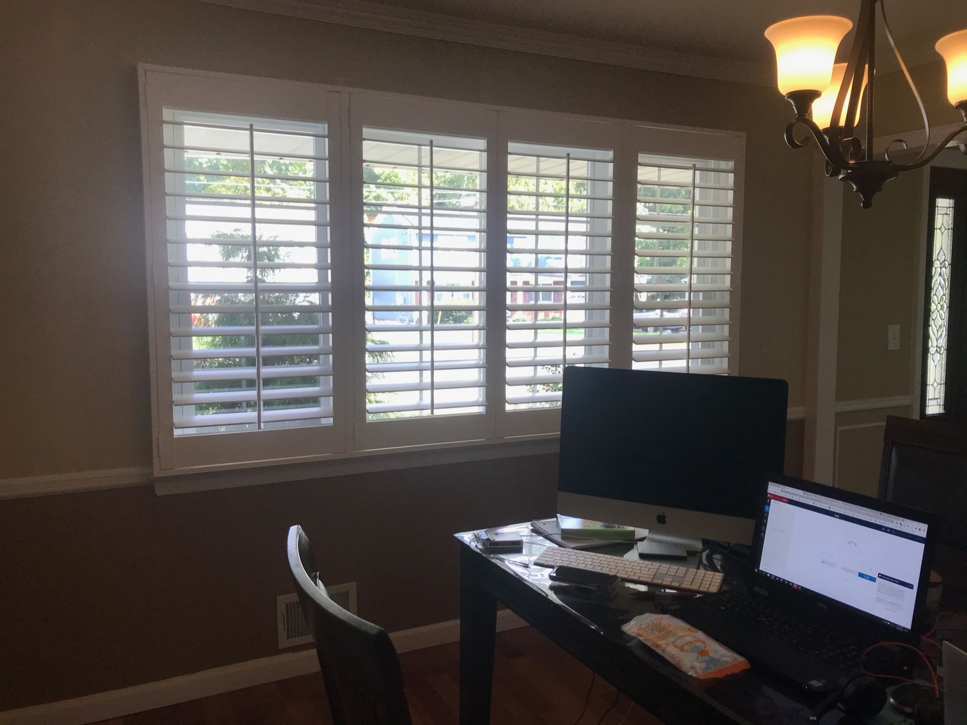 A window with white shutters behind a desk with computers and a chair.