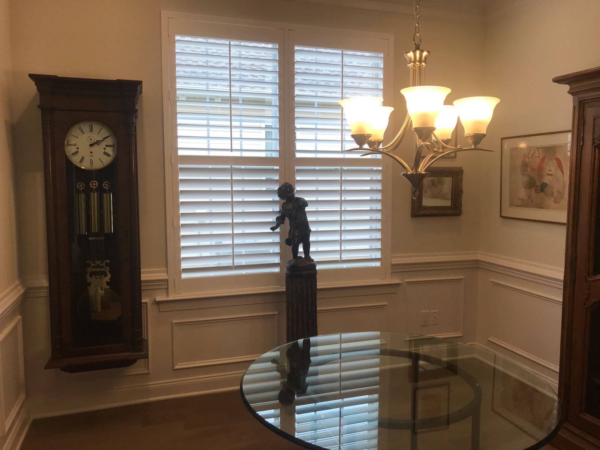 Dining room with glass table, window with shutters, grandfather clock, and a chandelier.