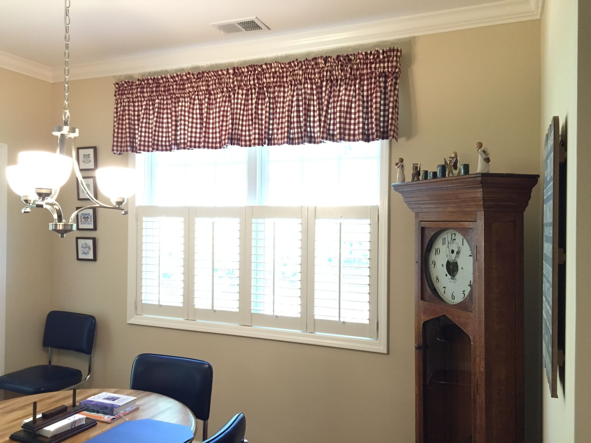 Window with red checked valance, white shutters, grandfather clock, and dining table.