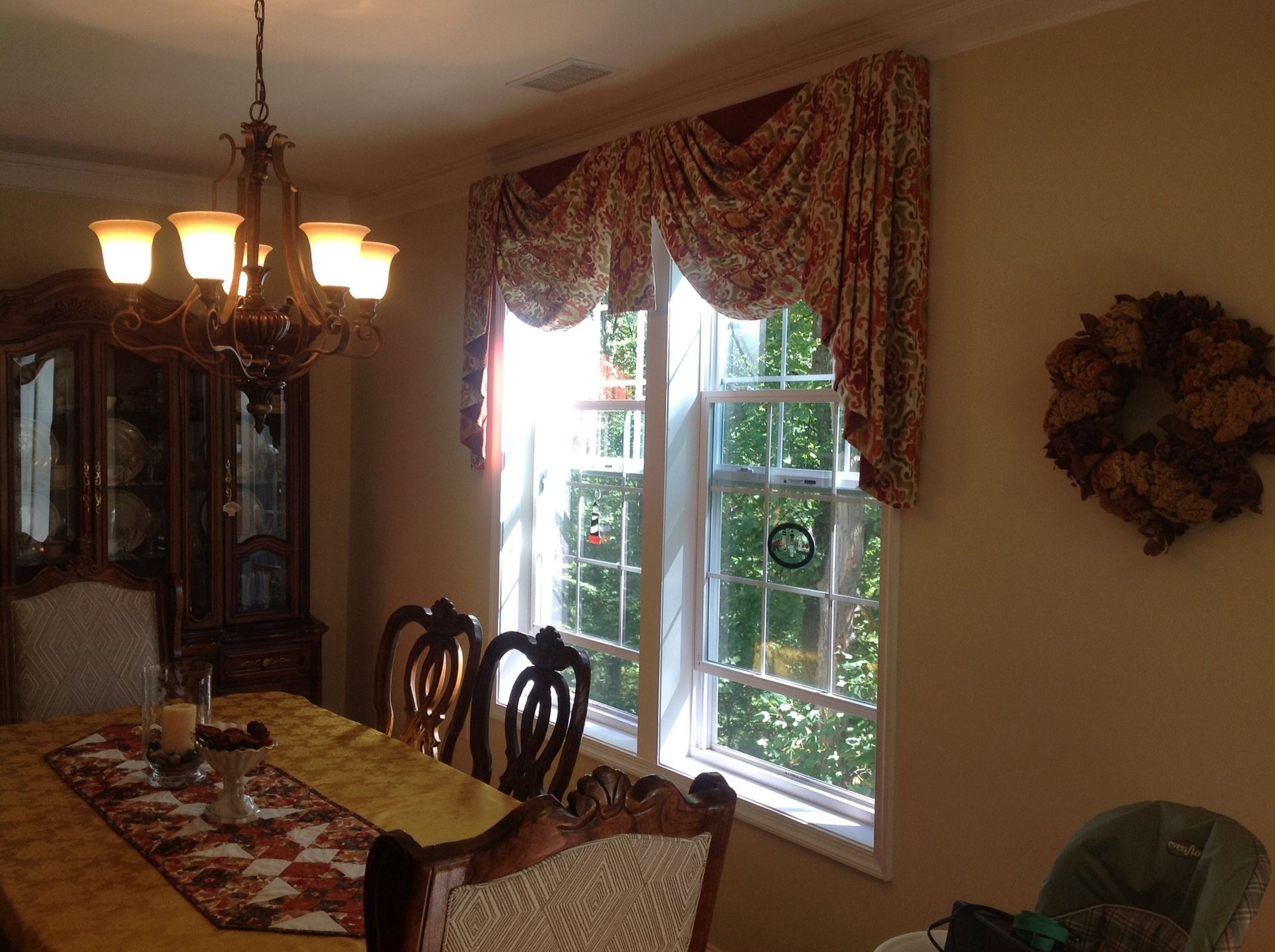 Dining room with table set for a meal; window with ornate draping; chandelier, buffet, and wreath.