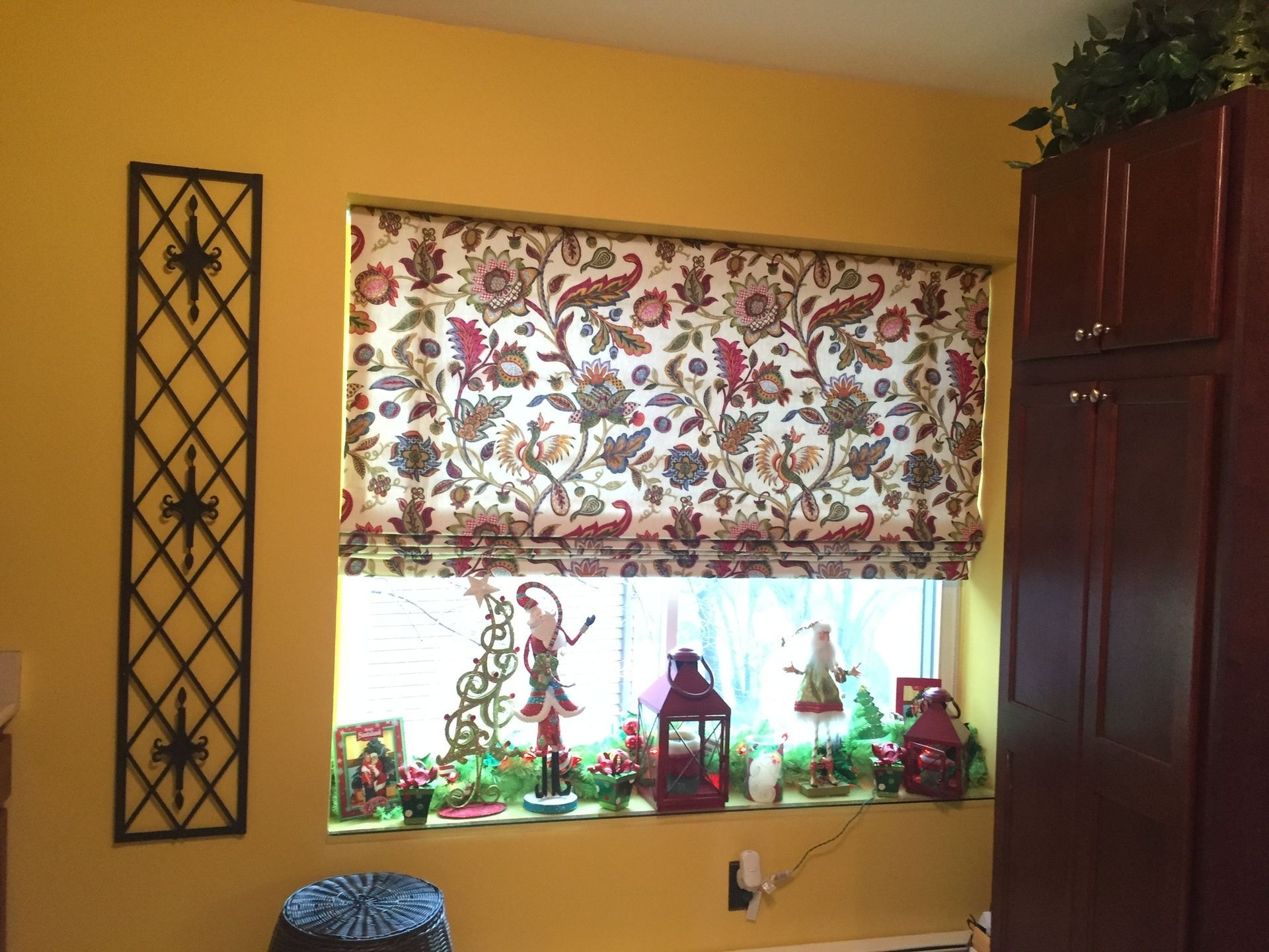 Window with floral Roman shade, Christmas decor on sill, yellow wall, and dark cabinet.