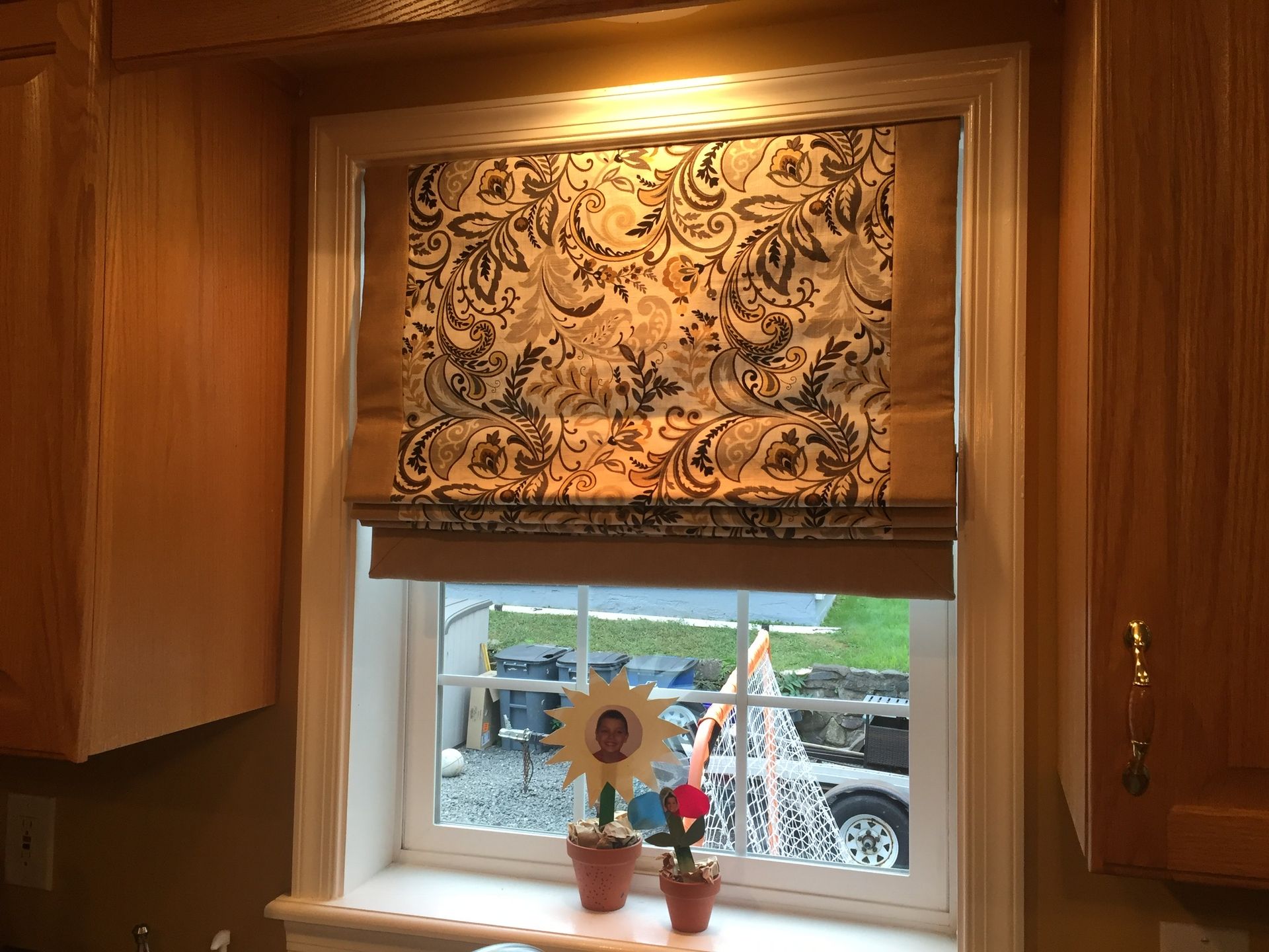 Window with floral Roman shade, framed by wooden cabinets. Sunlit, with view of garden.