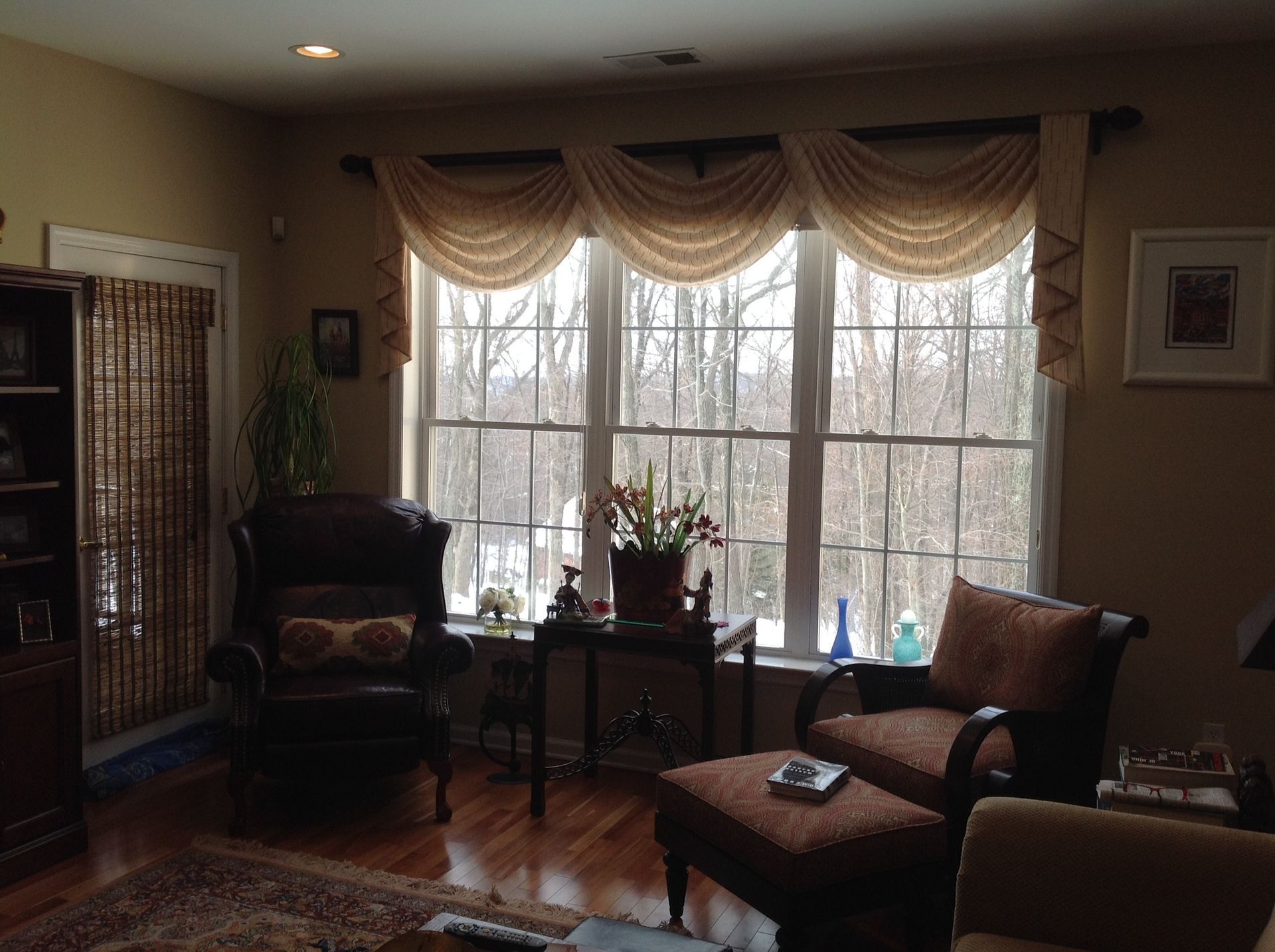 Cozy living room with a large window overlooking a forest, furnished with chairs, an ottoman, and a bookshelf.