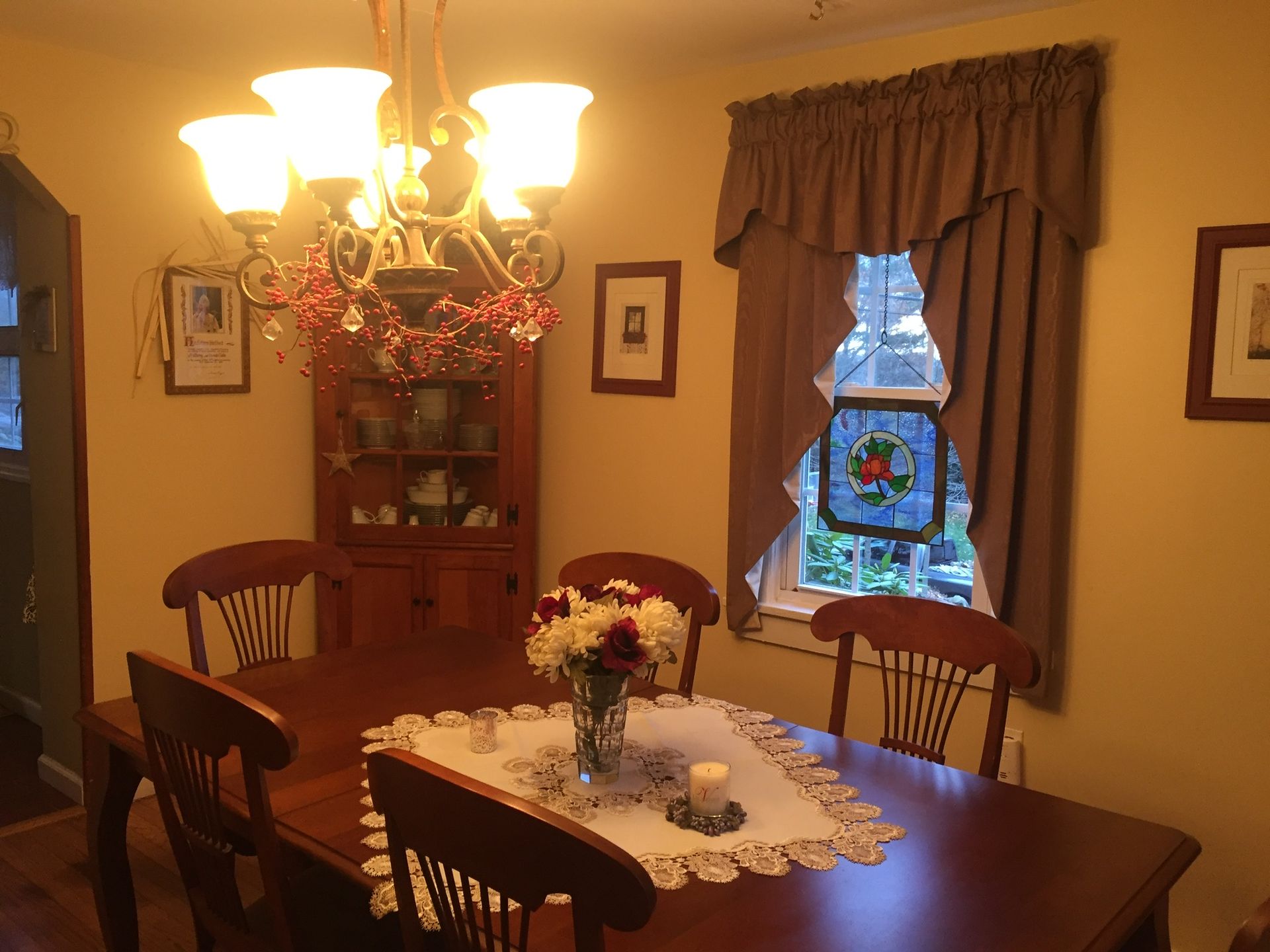 Dining room with wooden table, chairs, chandelier, corner cabinet, and window with curtains.