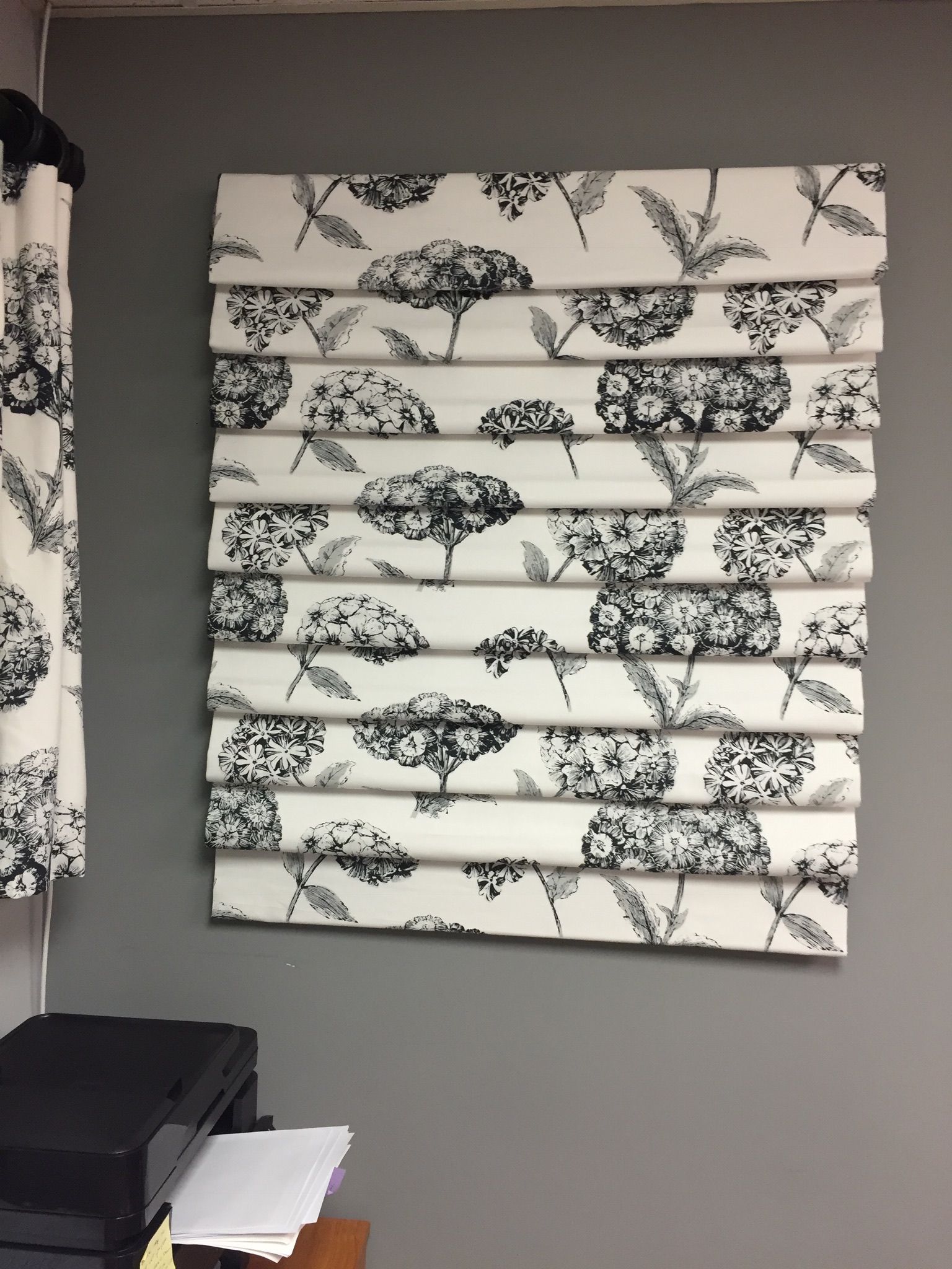 White and black floral Roman shade on a gray wall, next to matching curtains.