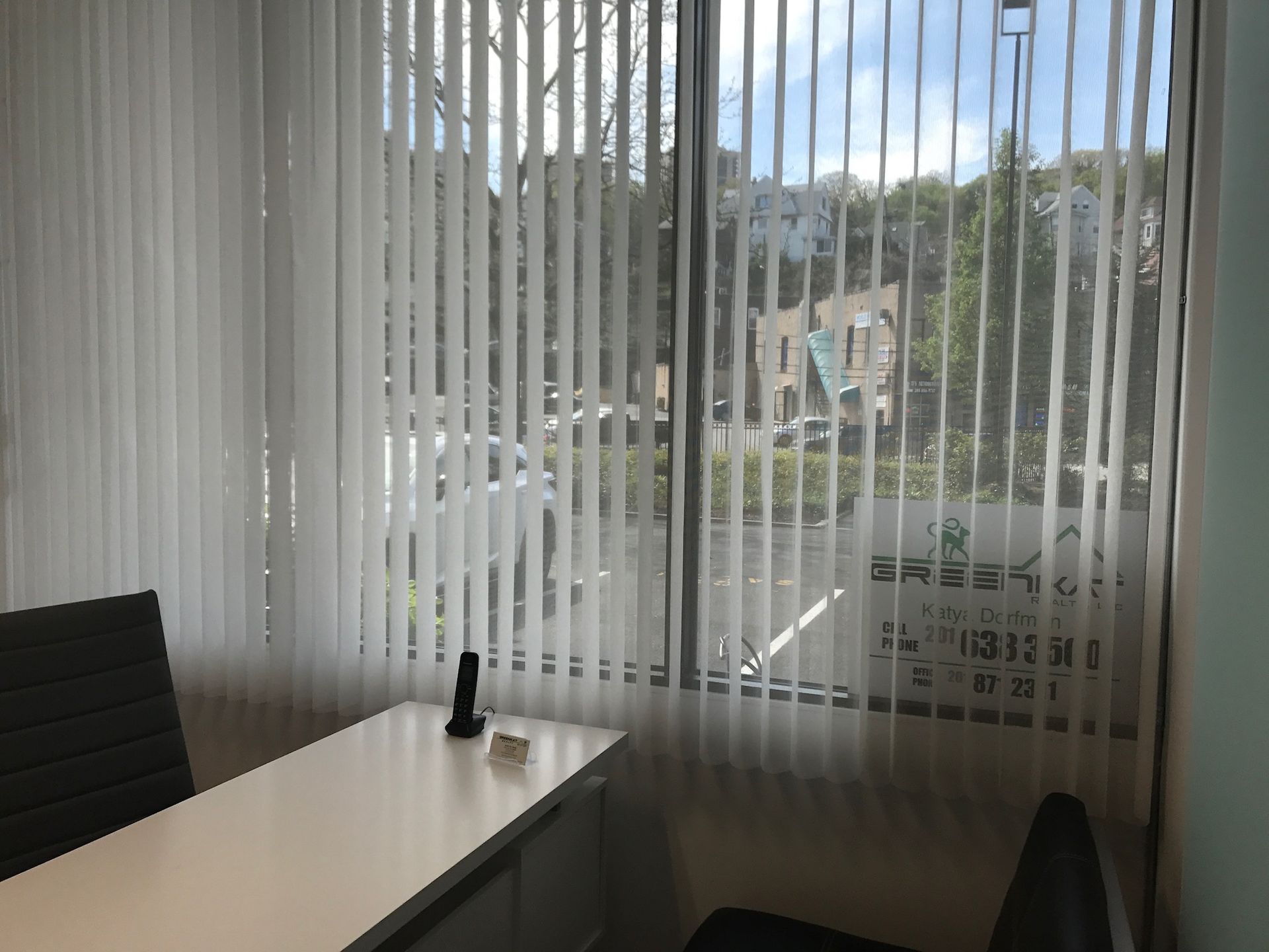 Office interior with a window, vertical blinds, desk, and chairs. View outside shows trees and buildings.