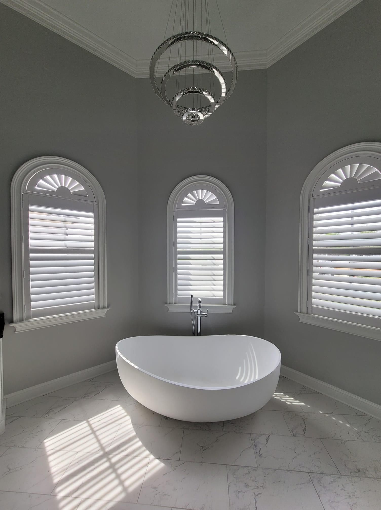 Elegant gray bathroom with a white tub beneath arched windows and a chandelier.