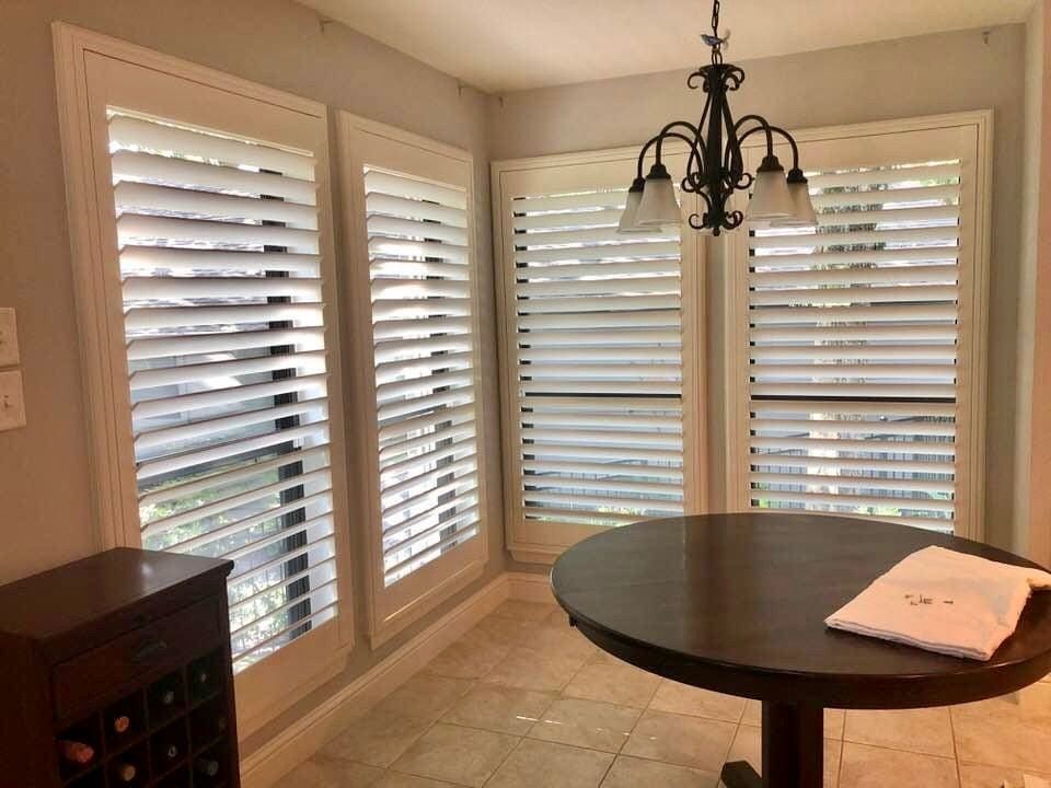 A sunlit breakfast nook with white shutters, round dark wood table, and a small chandelier.