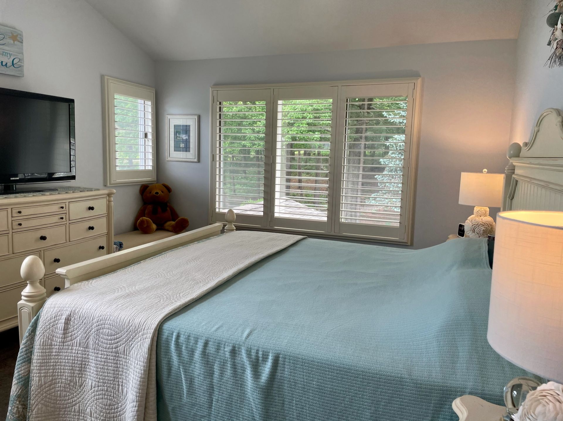 Bedroom with a bed, dresser, TV, and windows with shutters. Light blue bedspread, teddy bear, and lamps.
