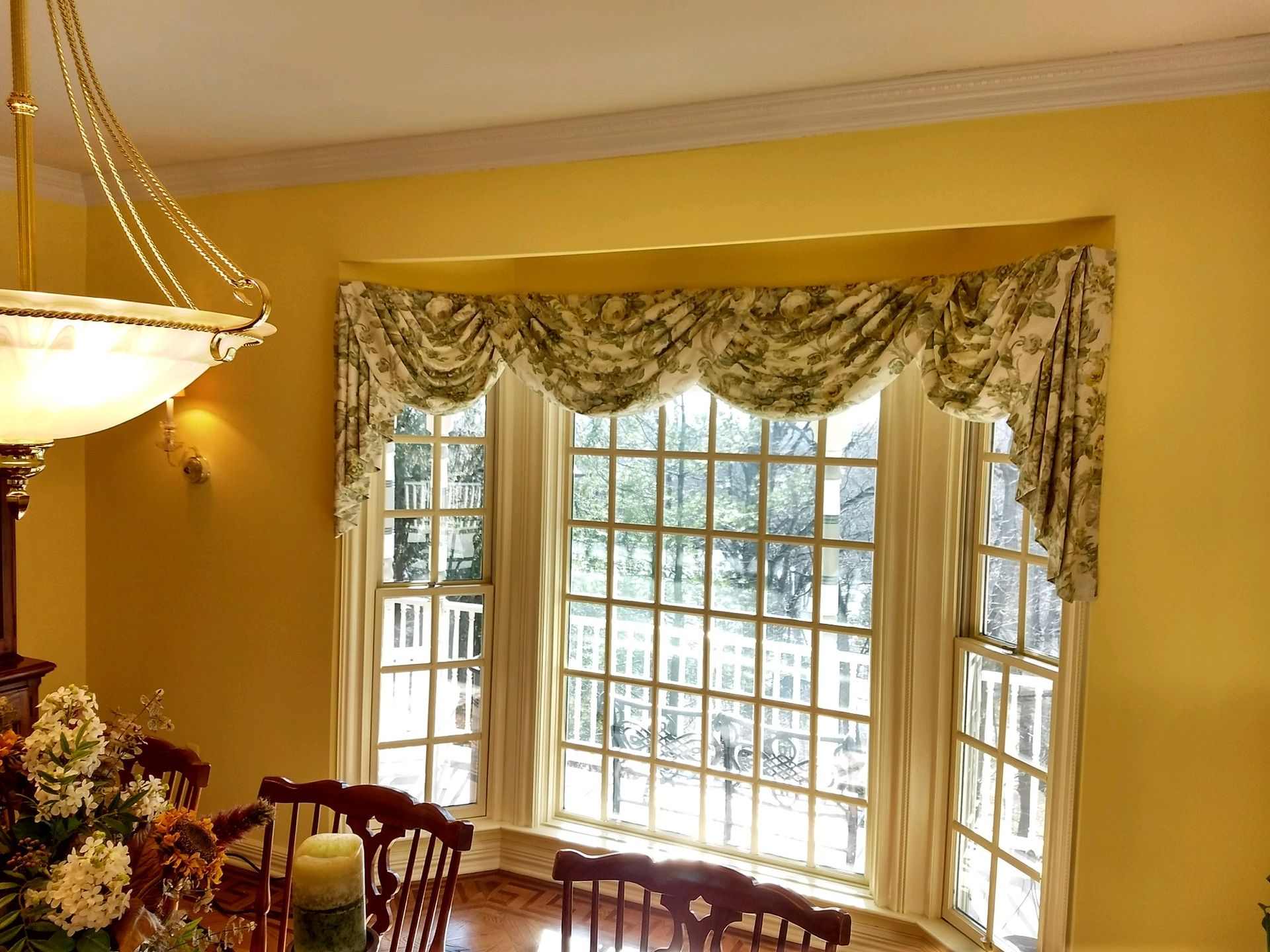 Bay window with floral valance, cream curtains, and yellow walls; interior dining room.