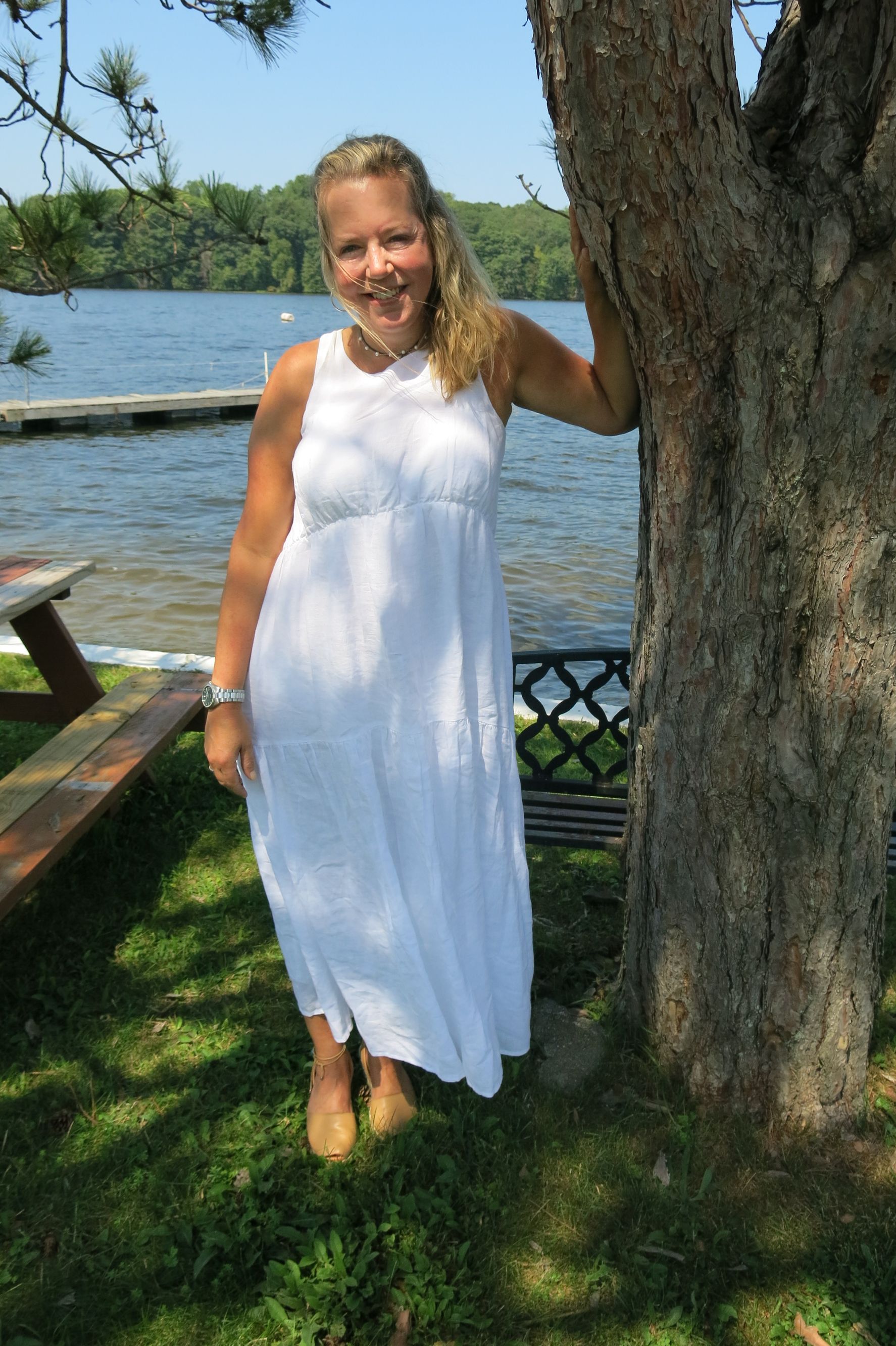 Woman in white dress smiles, leaning against a tree by a lake.