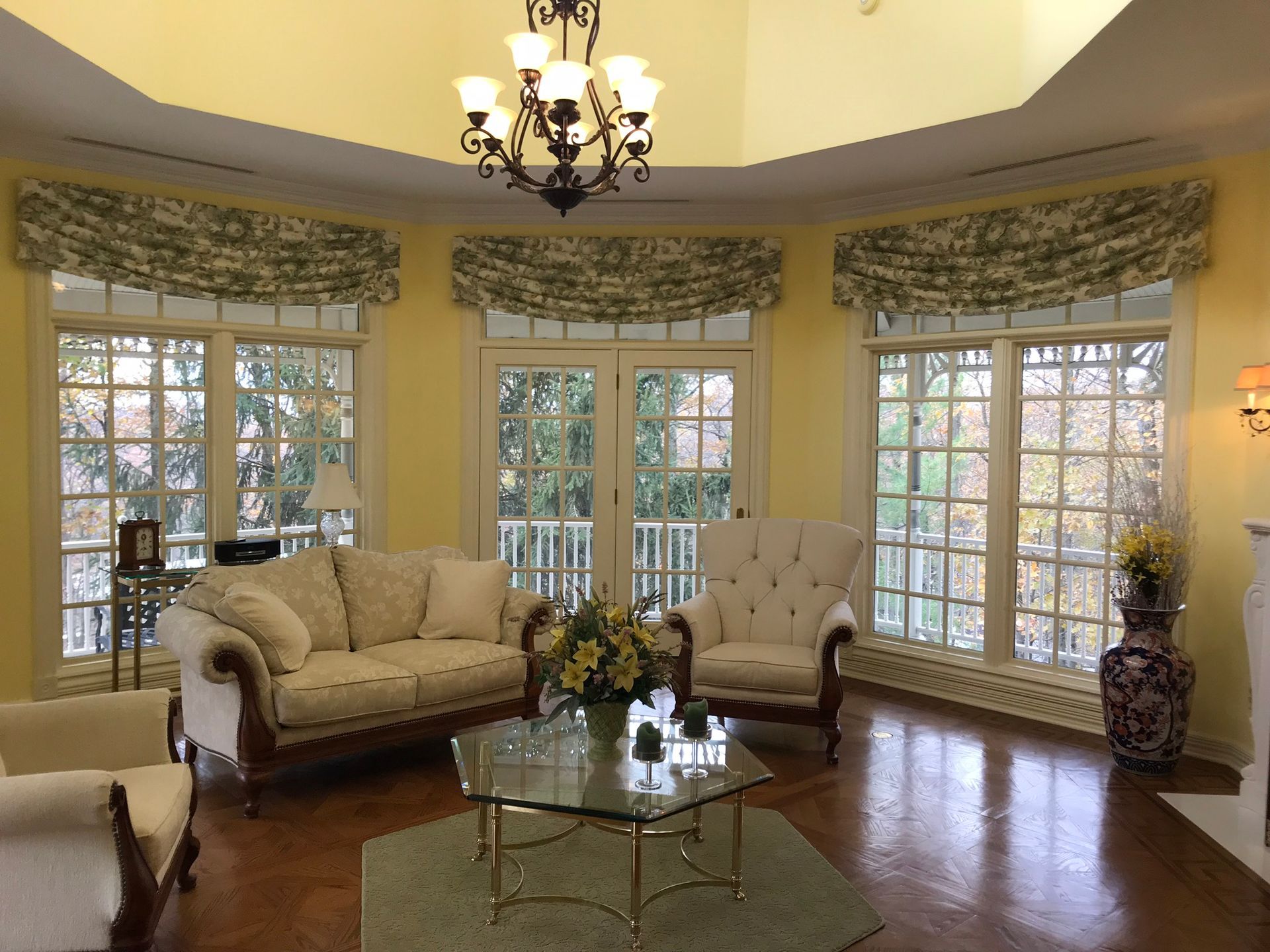 A living room with large windows, a couch, and chairs. Yellow walls and floral curtains.