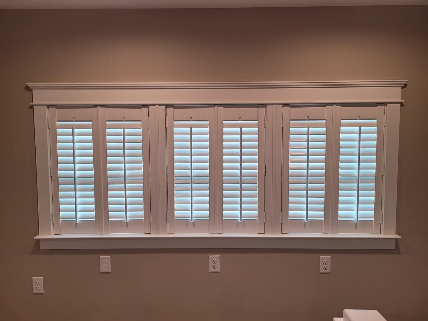 White shutters cover a window with three sections, mounted on a beige wall with white trim.
