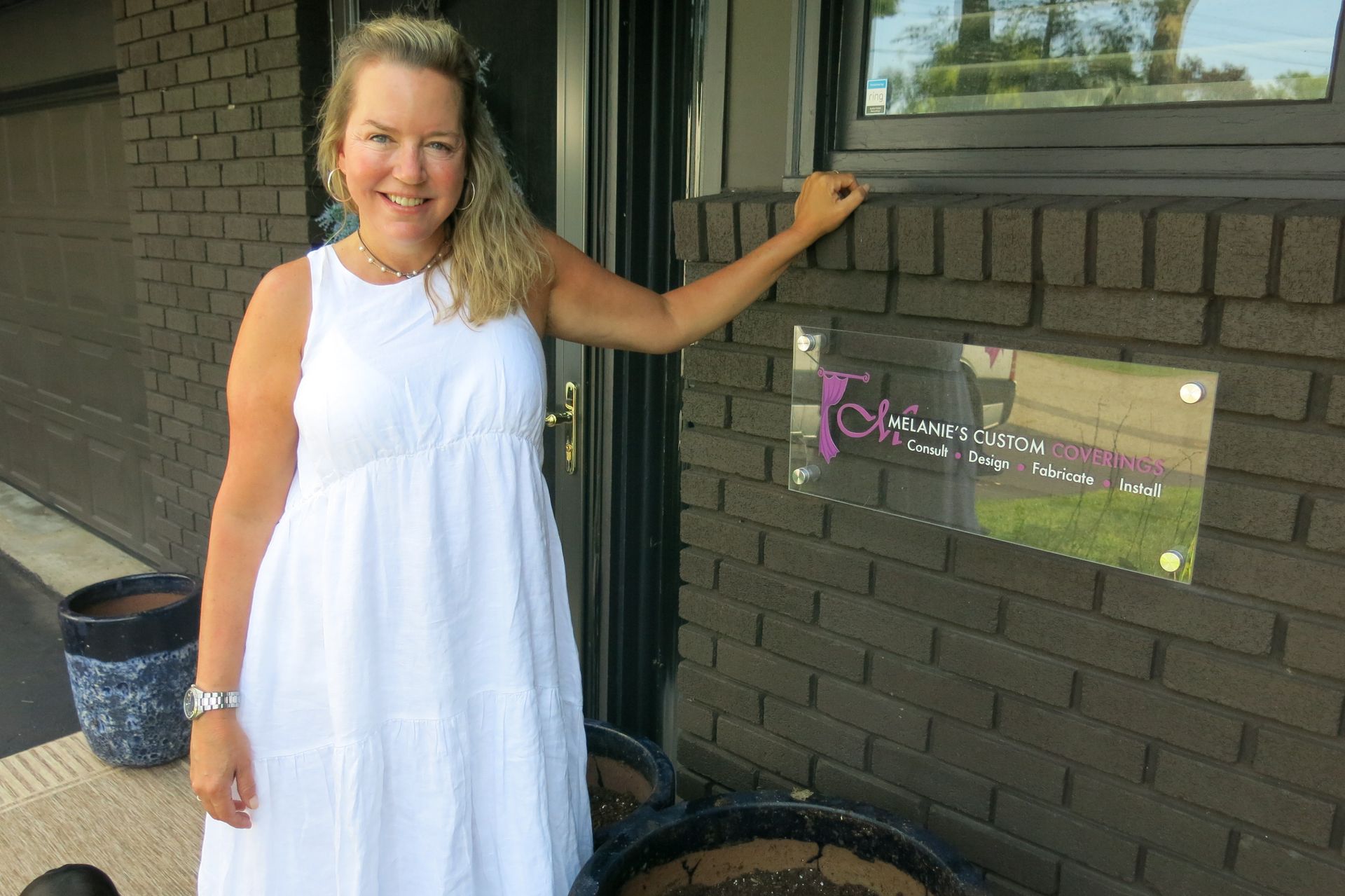 Woman in white dress smiles, pointing at a business sign on a brick building.