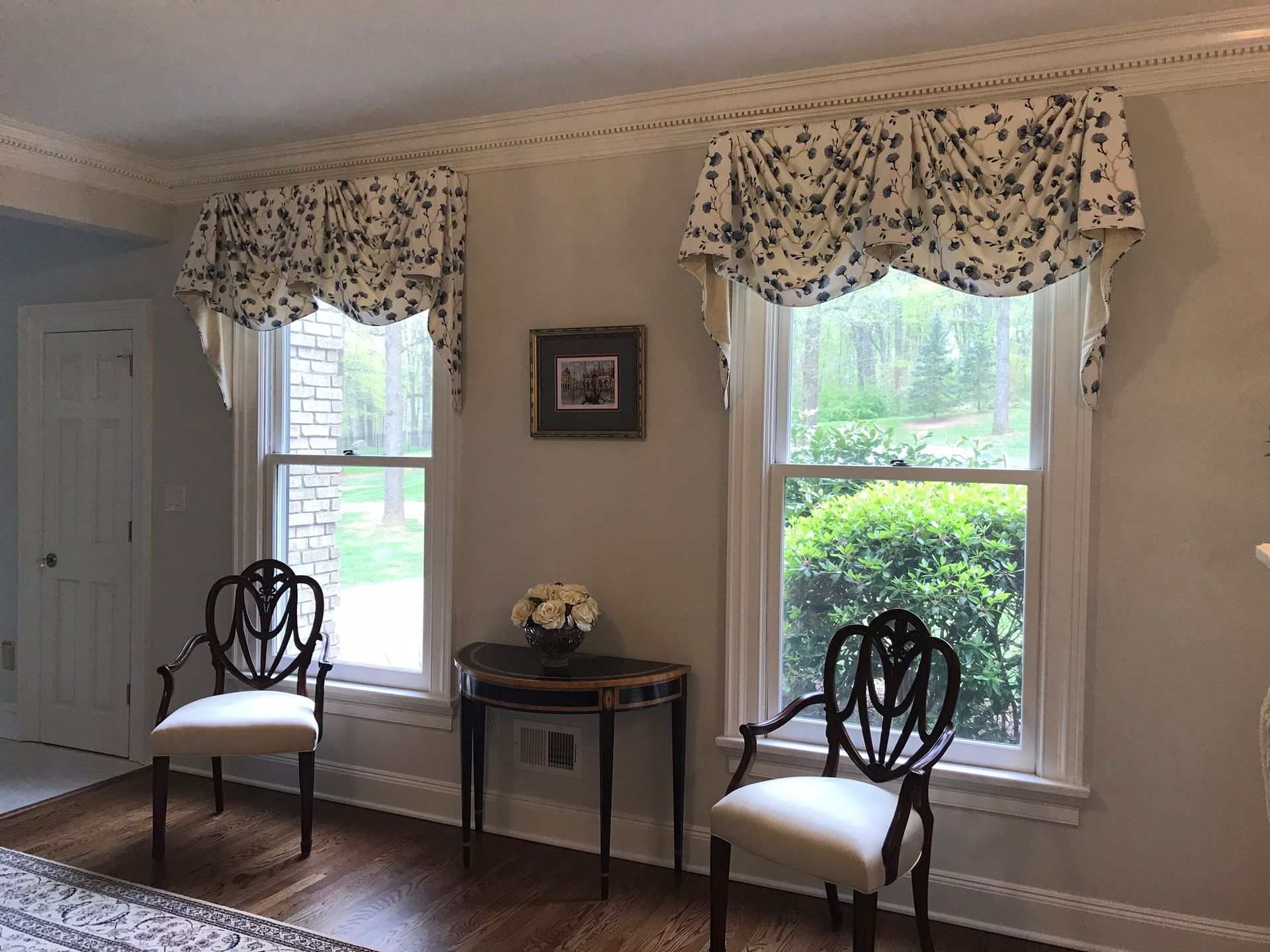 Two windows with floral valances in a room with antique chairs, table, and a picture.
