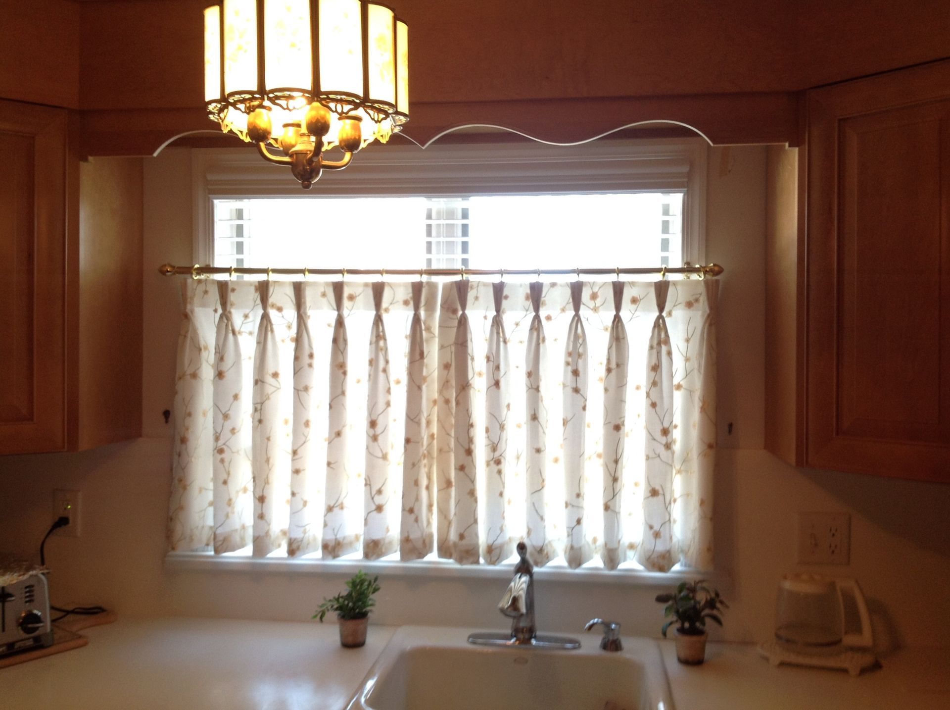Kitchen window with floral curtains, sink, overhead light, and light-colored cabinets.
