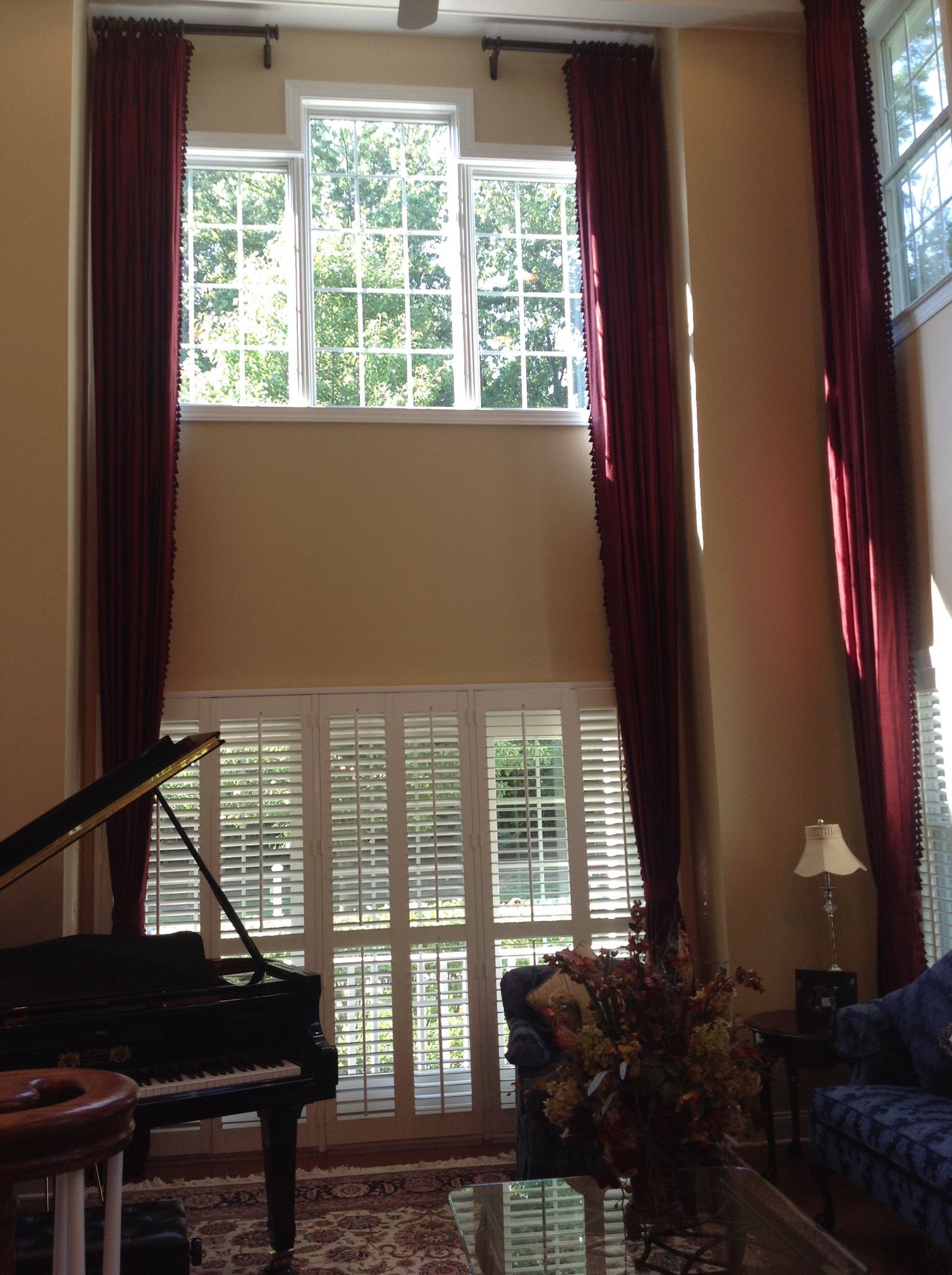 Living room with grand piano, two-story windows, red curtains, and a small seating area.