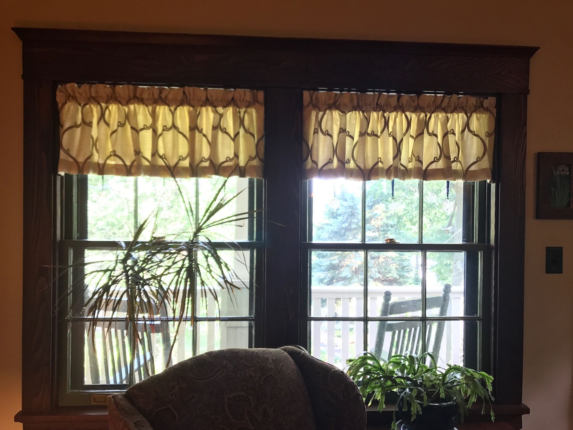 Double-hung windows with tan and brown valances and a dark wooden frame; a chair and plants are visible.