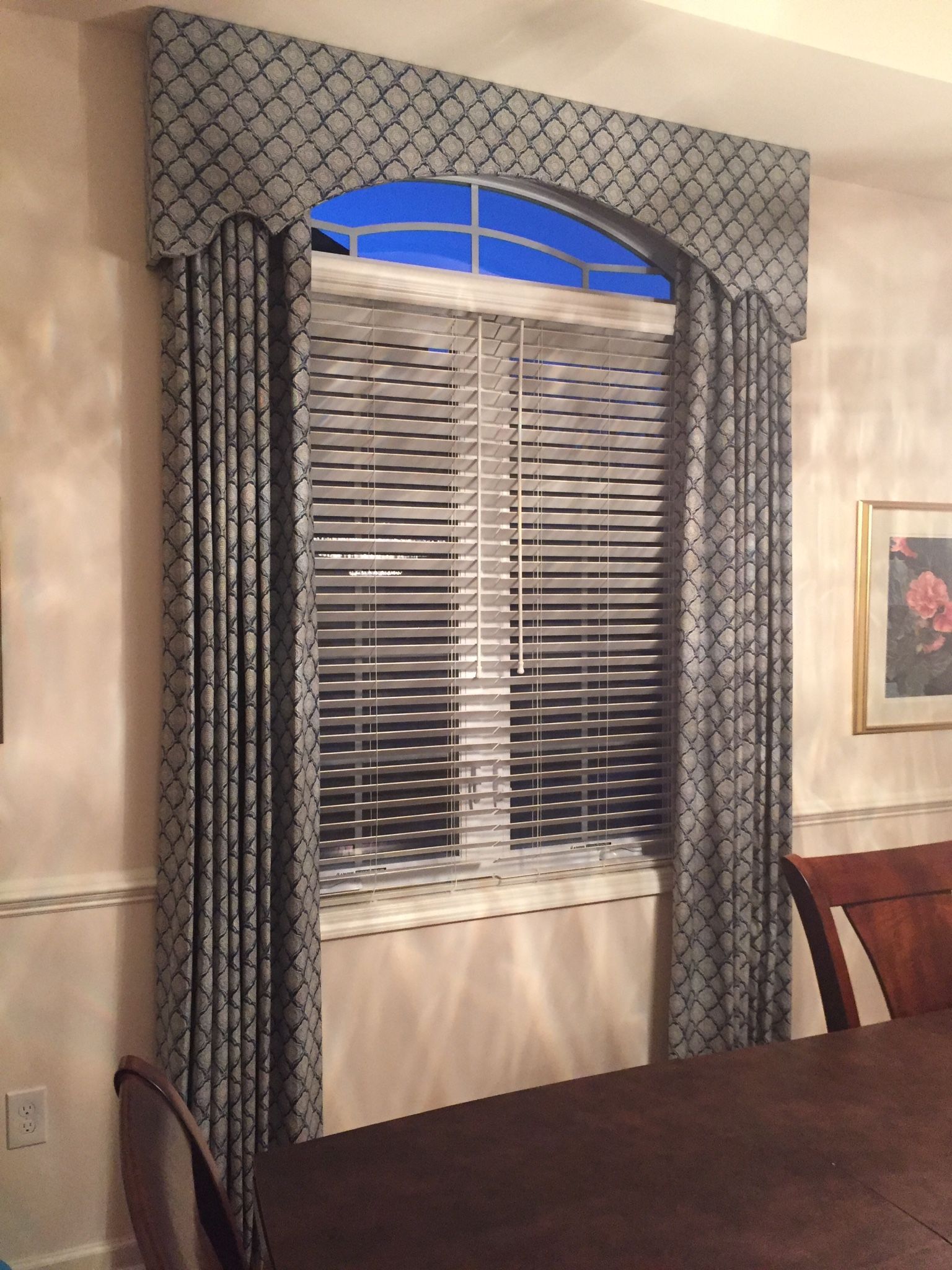 Window with gray patterned valance and curtains framing blinds, a dark table and chair are in the foreground.