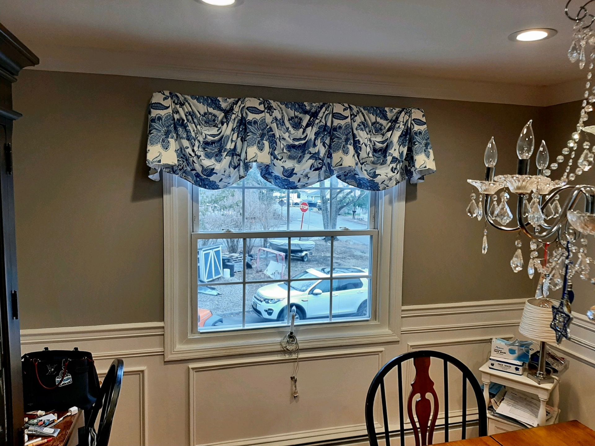 A window with a blue and white valance above it, with a view of outside, and part of a chandelier.