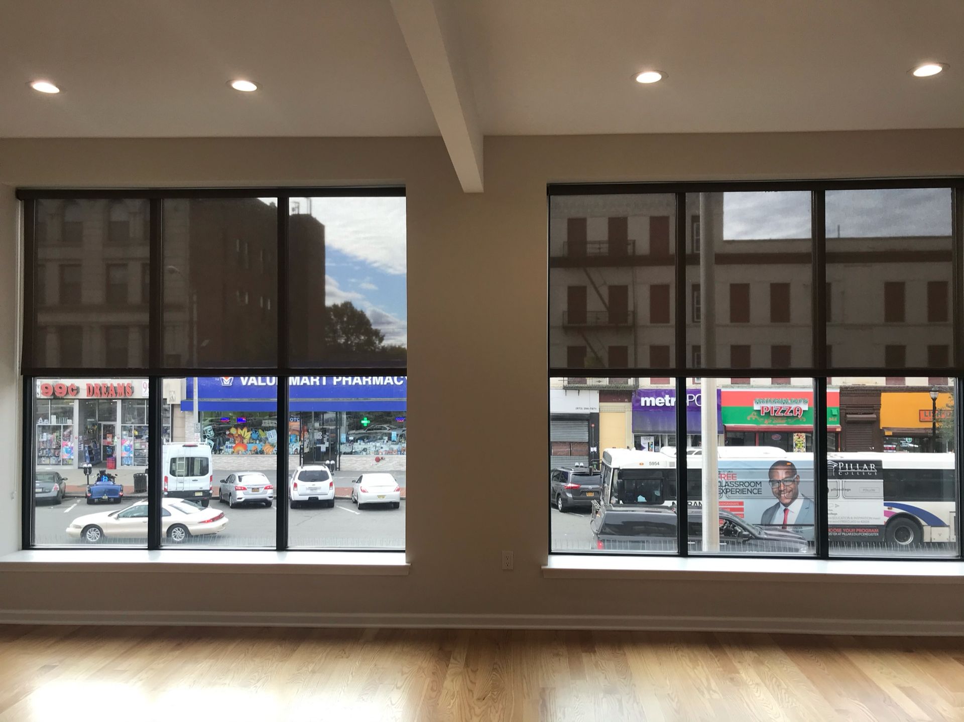 Two large windows with black blinds, looking out onto a city street with cars and buildings.