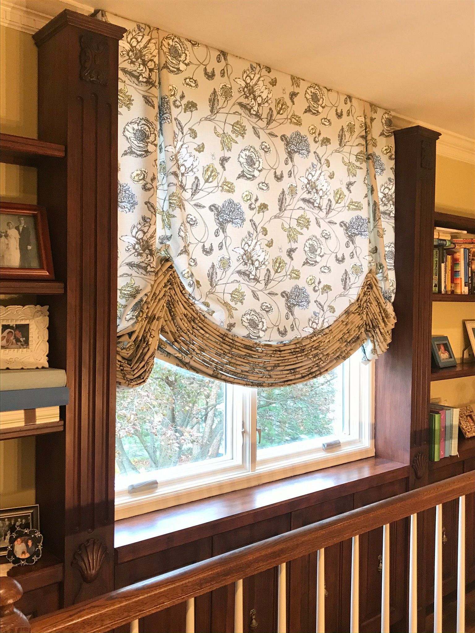 Roman shade with floral pattern, brown trim, over a window in a room with built-in bookshelves and a railing.