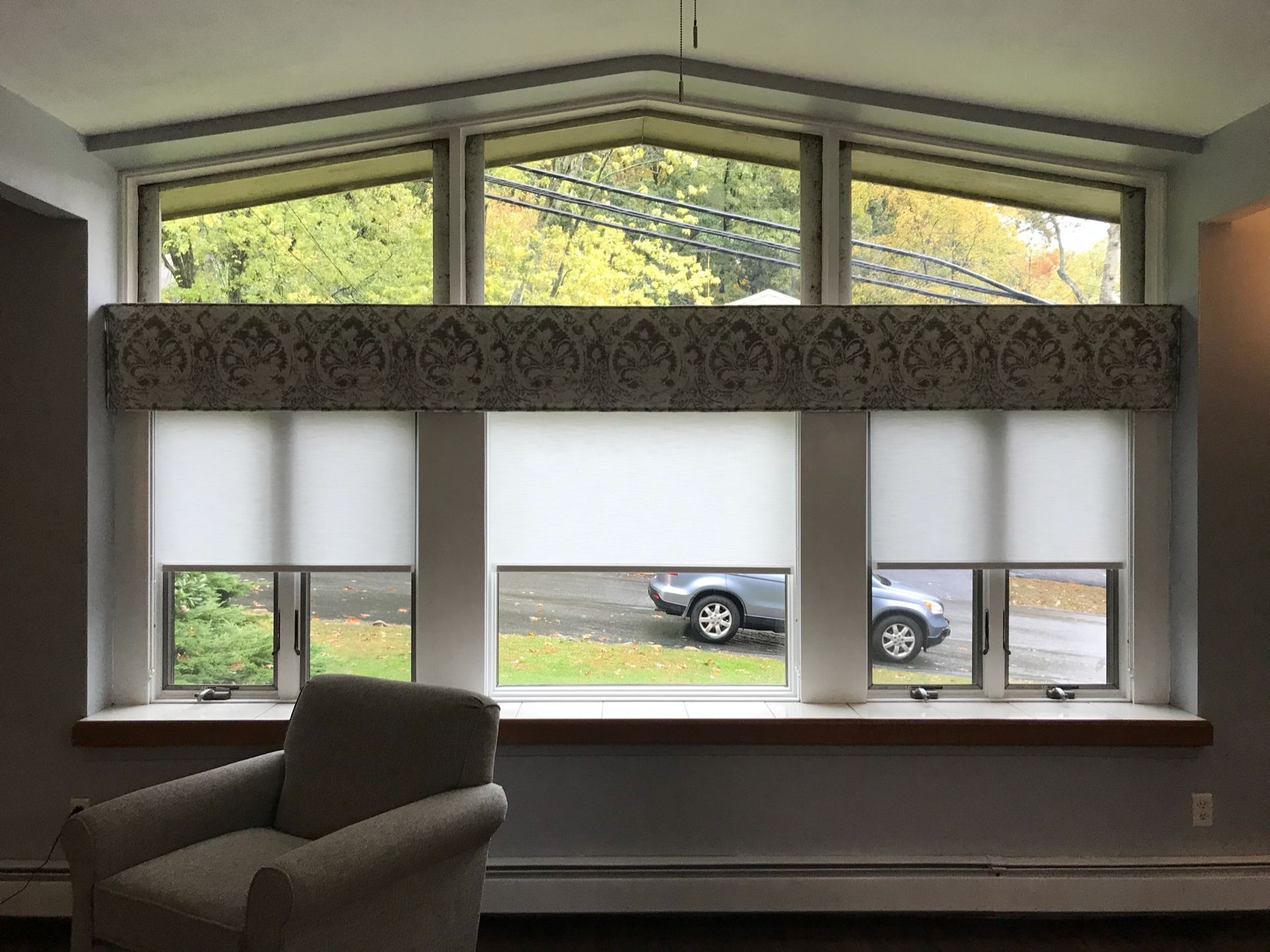 A room with a large window featuring three white roller blinds, topped with a patterned valance. A car is visible outside.
