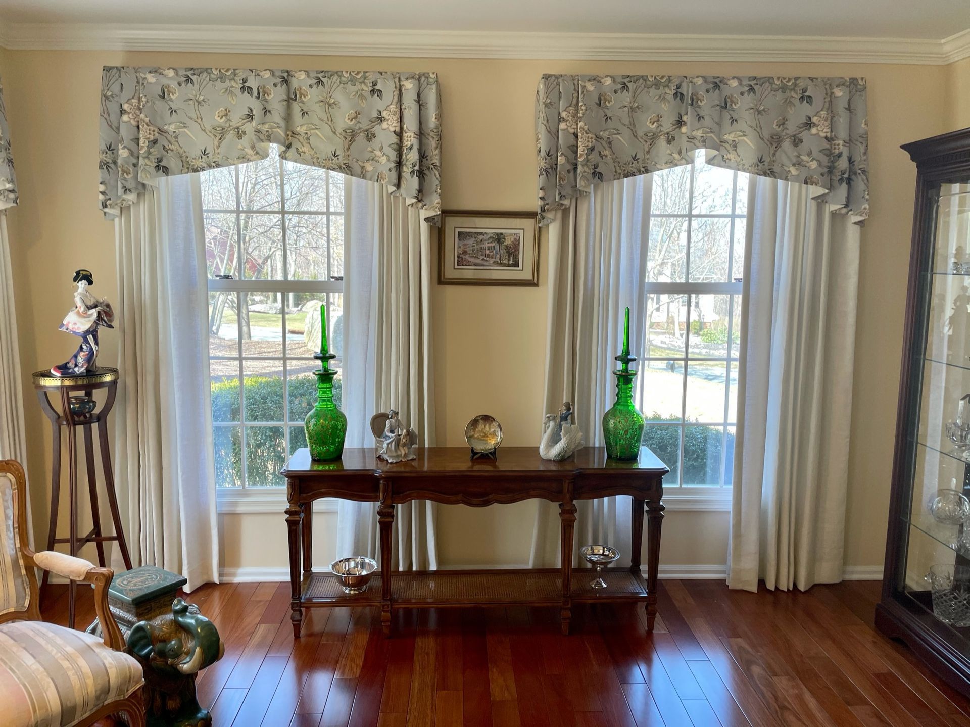Two windows with floral valances and cream curtains, a dark wood table with decor, and a glass cabinet in a room.