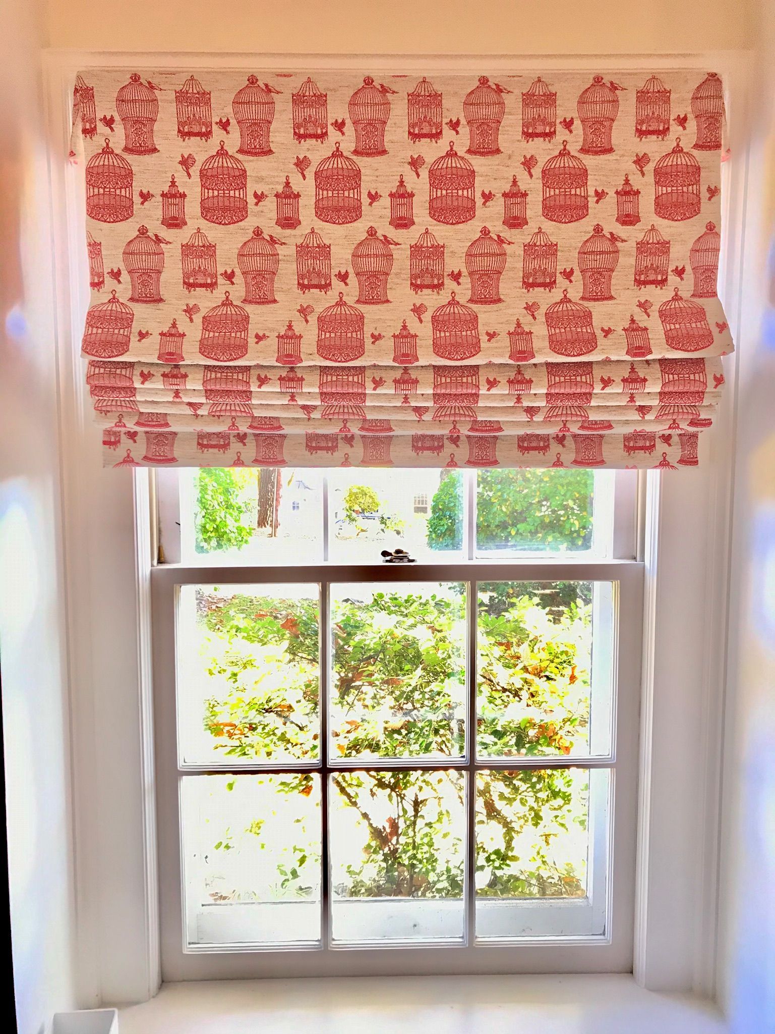 Red patterned roman shade covering a white window. Bright sunlight shines through.
