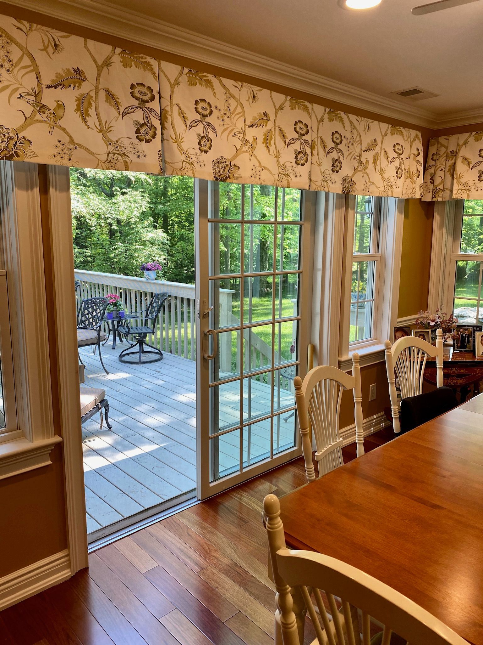 A dining room with a view of a deck, sliding glass door, floral valance, and a wooden table.