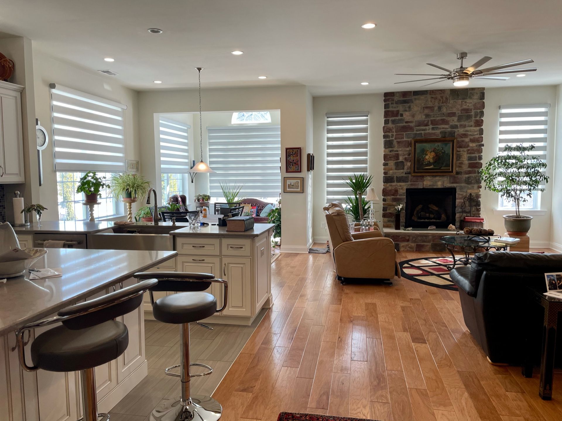 Open-concept living area with kitchen island, stone fireplace, wood floors, and large windows with blinds.