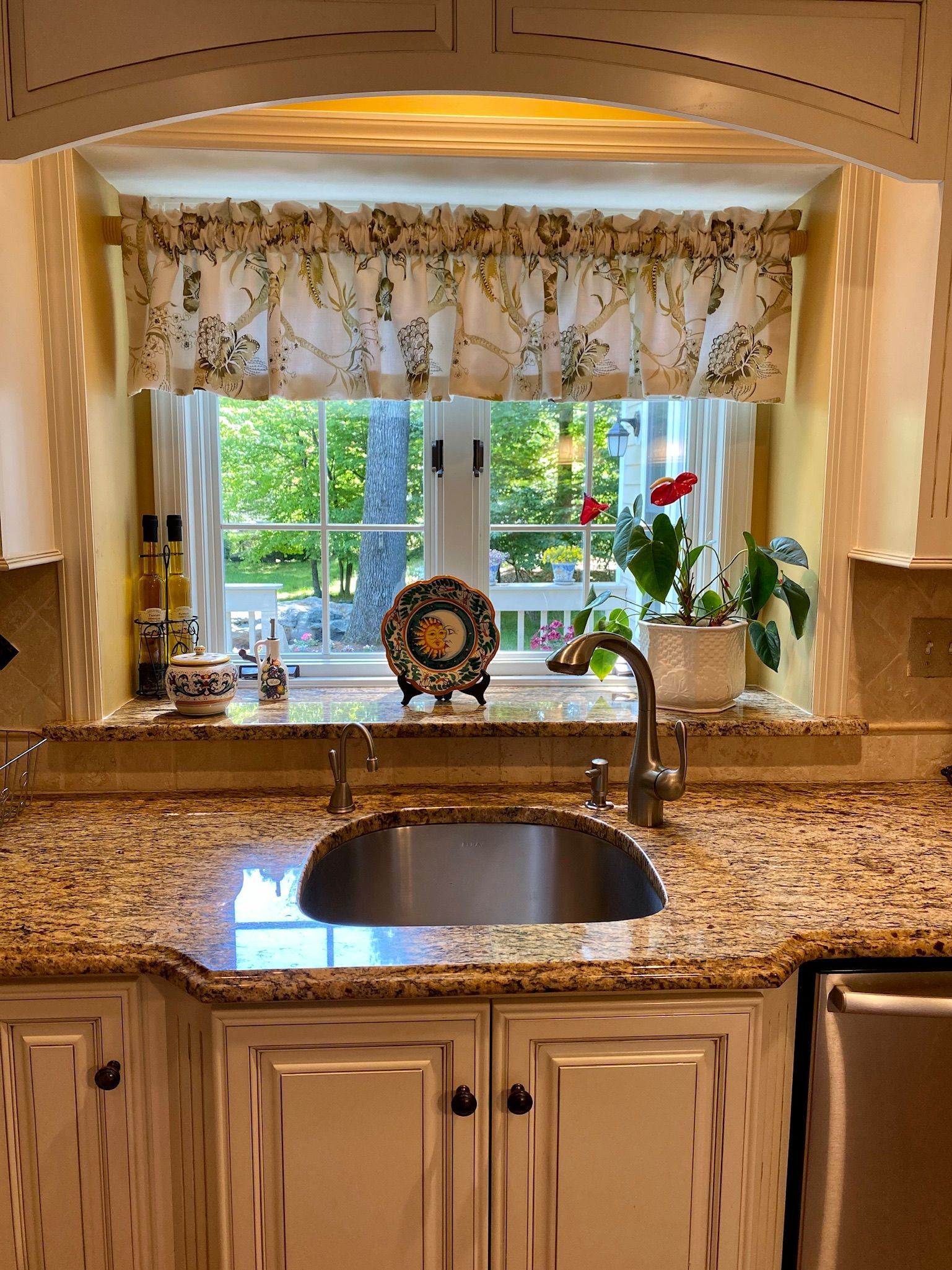 Kitchen sink area with granite countertops, white cabinets, and a window with a floral valance overlooking a green outdoor view.