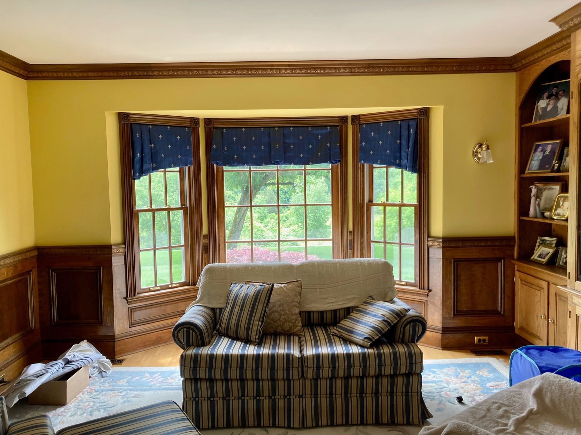 Yellow-walled room with wood trim, a striped sofa, and a window with blue valances overlooking a green yard.