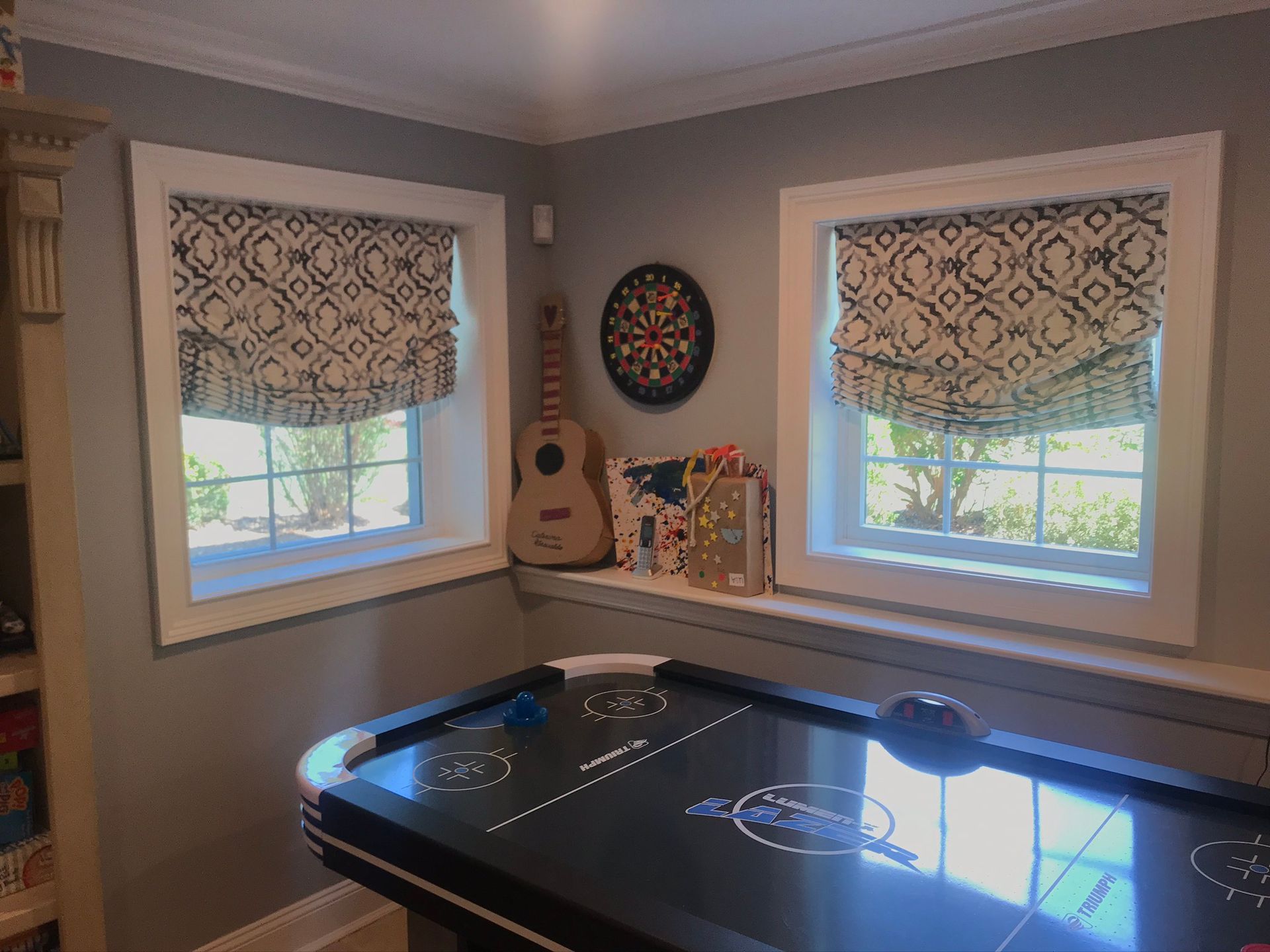 Game room with air hockey table, guitar, dartboard, and windows with patterned shades.