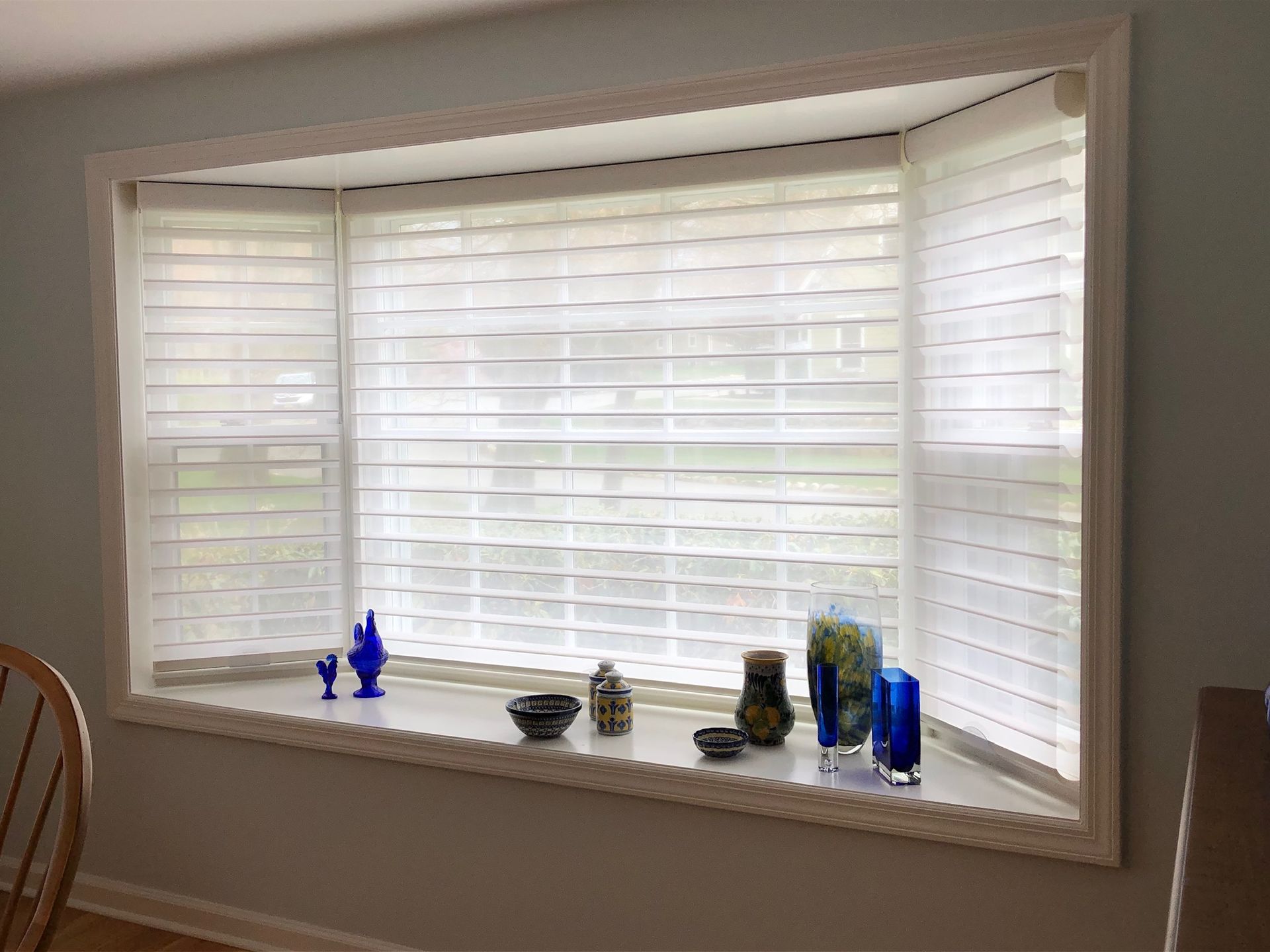 Bay window with white blinds, decorative glass pieces on the sill, and a chair in view.