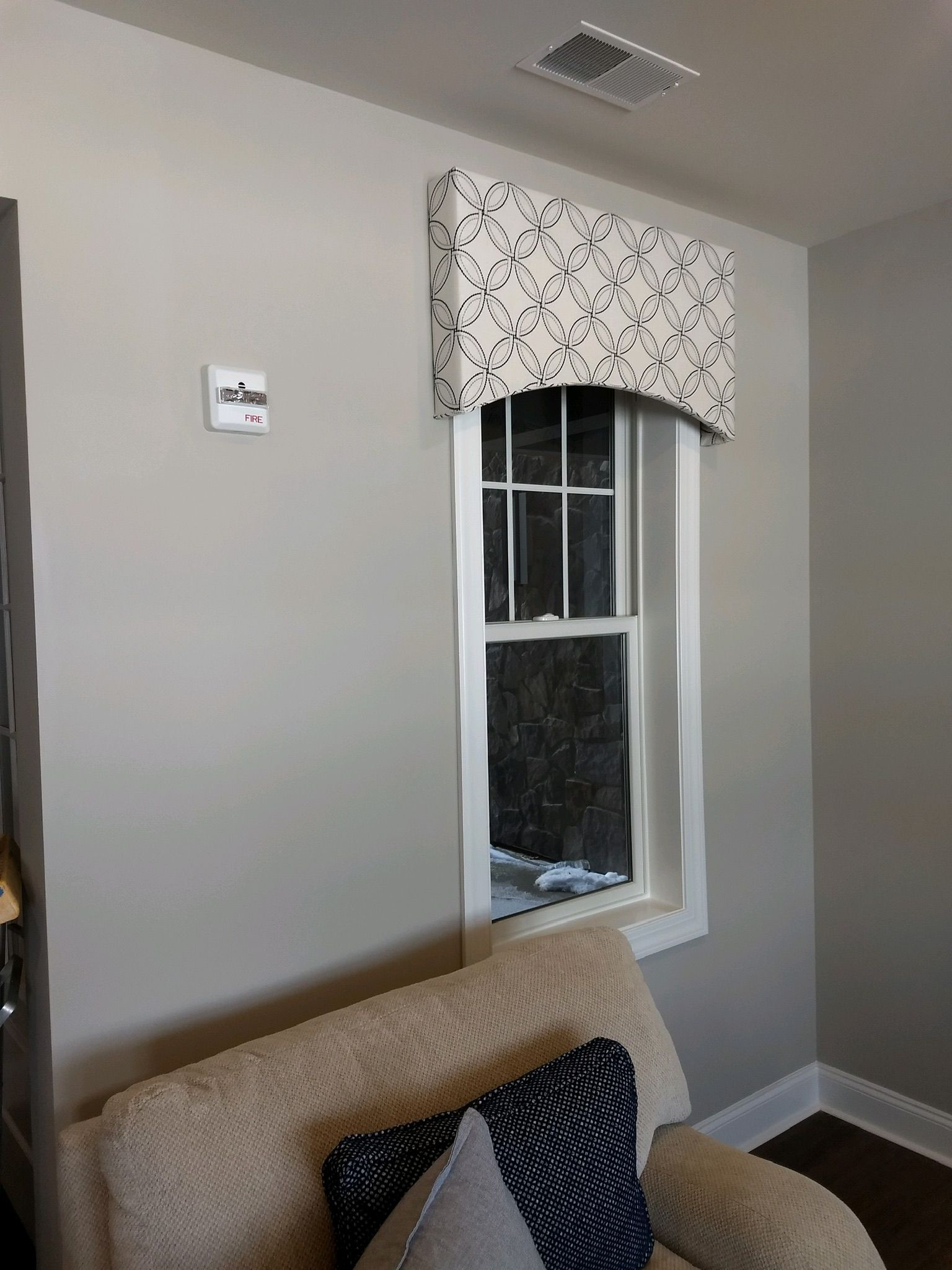 Window with a patterned valance above an armchair with decorative pillows, set in a room with a light gray wall.