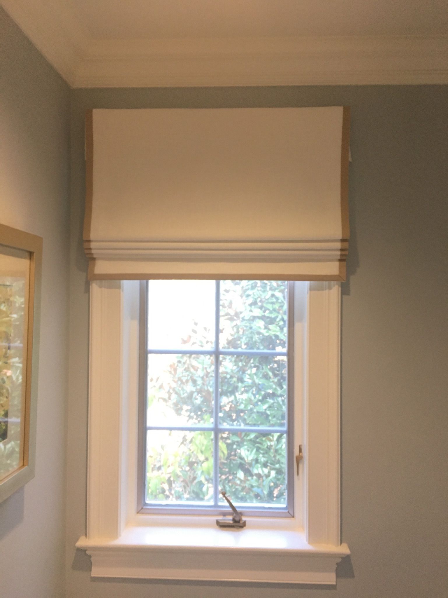 A window with a tan Roman shade, set in a white frame on a pale blue wall.