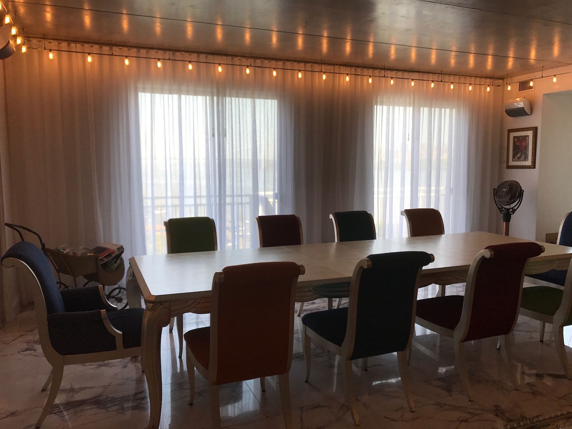 Dining room with long table and colorful chairs, sheer curtains, and string lights.